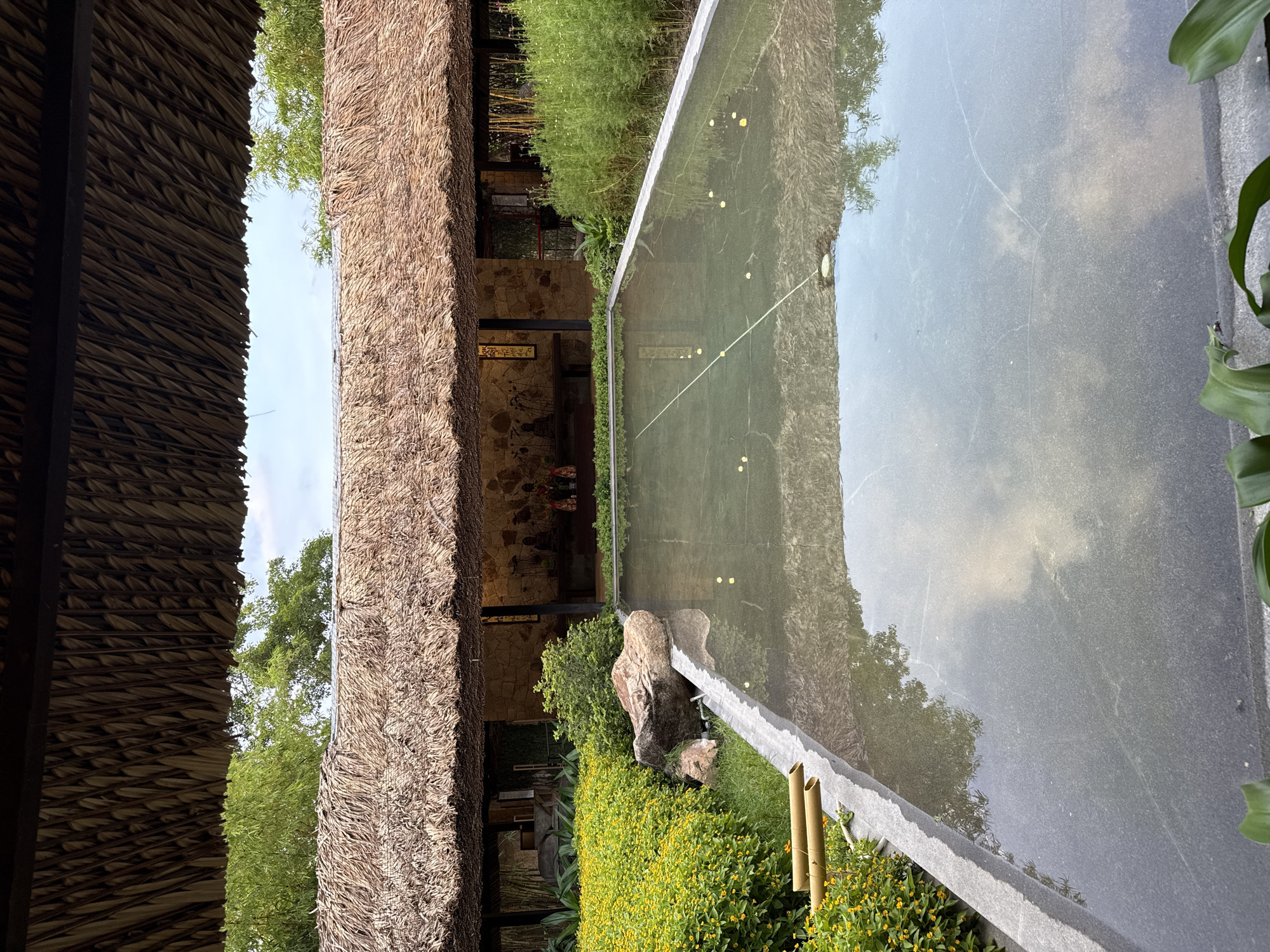 This outdoor scene in Tay Nha Trang Ward, Vietnam, shows a building with a thatched roof adjacent to a rectangular pool of water, reflecting the surrounding trees and sky. A stone wall is partially visible behind the thatched roof. The ground beside the pool is covered with green foliage and small yellow flowers. A large rock sits amidst the greenery. Two vertical bamboo poles stand near the edge of the pool. The overall impression is of a tranquil, landscaped environment.
