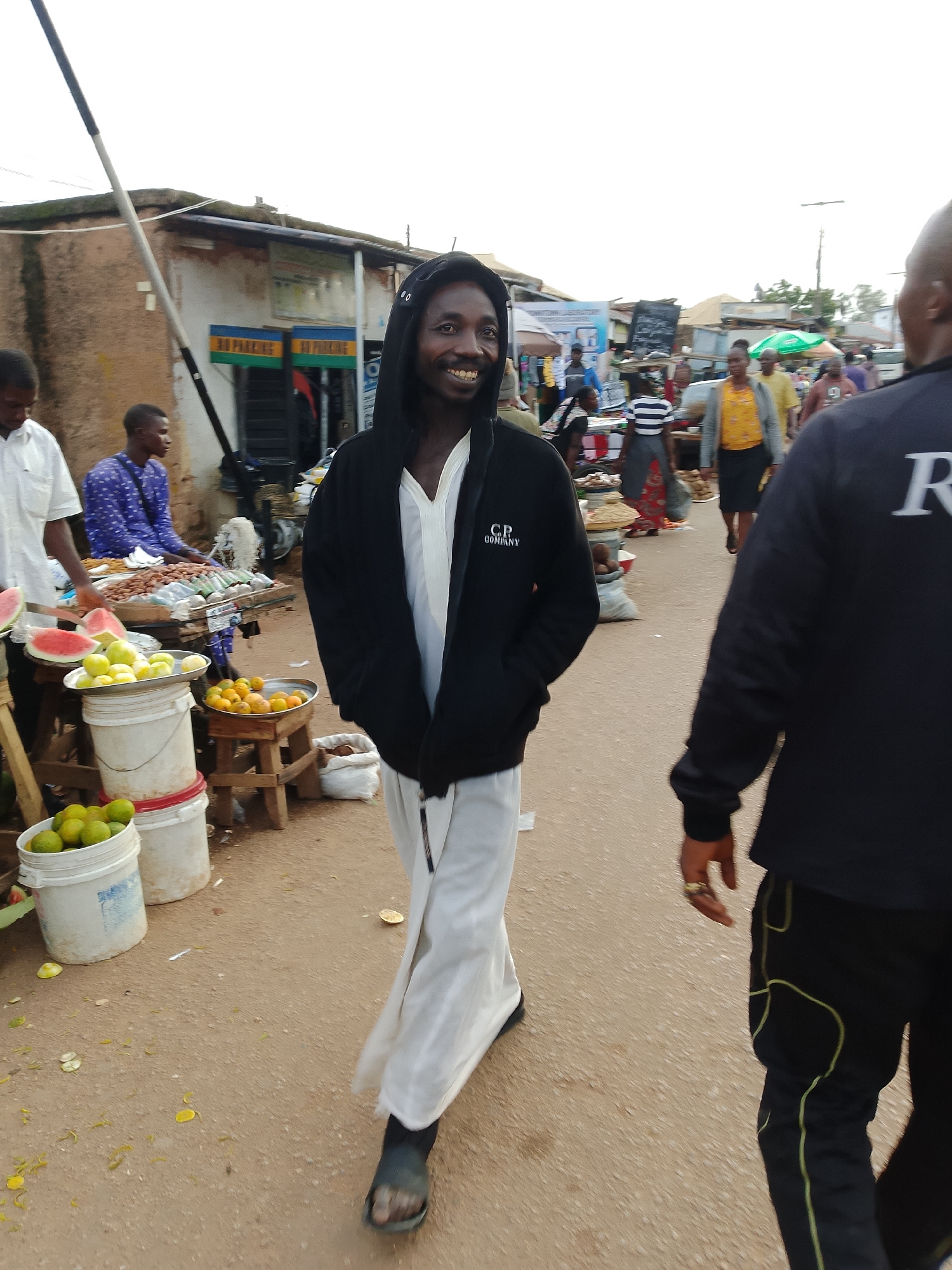 A man wearing a black hooded sweatshirt and white pants walks through a street market. He is smiling at the camera, and his hands are in his pockets. Behind him, other people are walking and shopping. There are stalls selling fruits and other goods. The ground is dirt and there are some fallen leaves.