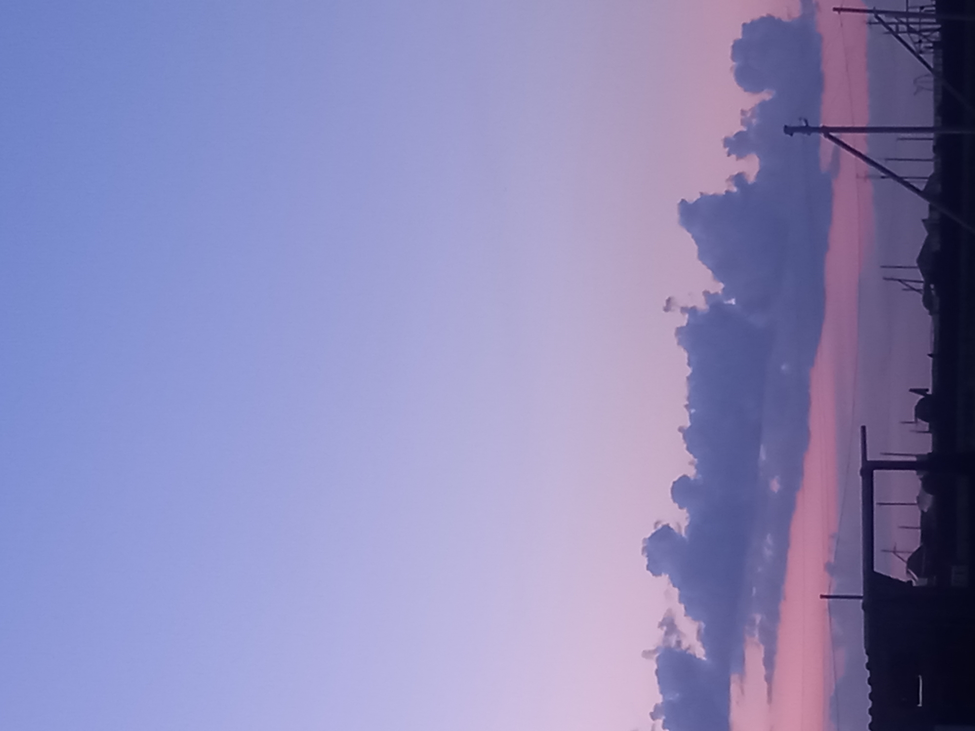 The image captures a twilight sky over an industrial or semi-rural area in Қозыкеткен ауылдық округі, Kazakhstan. The sky transitions from a pale lavender at the top to a soft pinkish hue near the horizon. A line of dark, irregularly shaped clouds stretches across the mid-ground. Below the clouds, silhouetted against the sky, are various structures. On the left, a dark, angular roofline of a building is visible. Towards the center and right, a series of utility poles, some with diagonal supports, are prominent, suggesting electrical infrastructure. The overall scene is characterized by muted colors and stark silhouettes, indicative of the fading light. No people are visible in the frame.