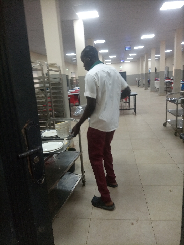 In a spacious hall with tiled floors and numerous pillars, a man stands near a utility cart piled with plates. He is dressed in maroon pants, a white shirt, and casual footwear. The scene suggests a dining or catering area, indicated by the arrangement of tables, chairs, and additional utility carts further down the hall. Bright ceiling lights illuminate the space. Part of a dark door is visible in the foreground.