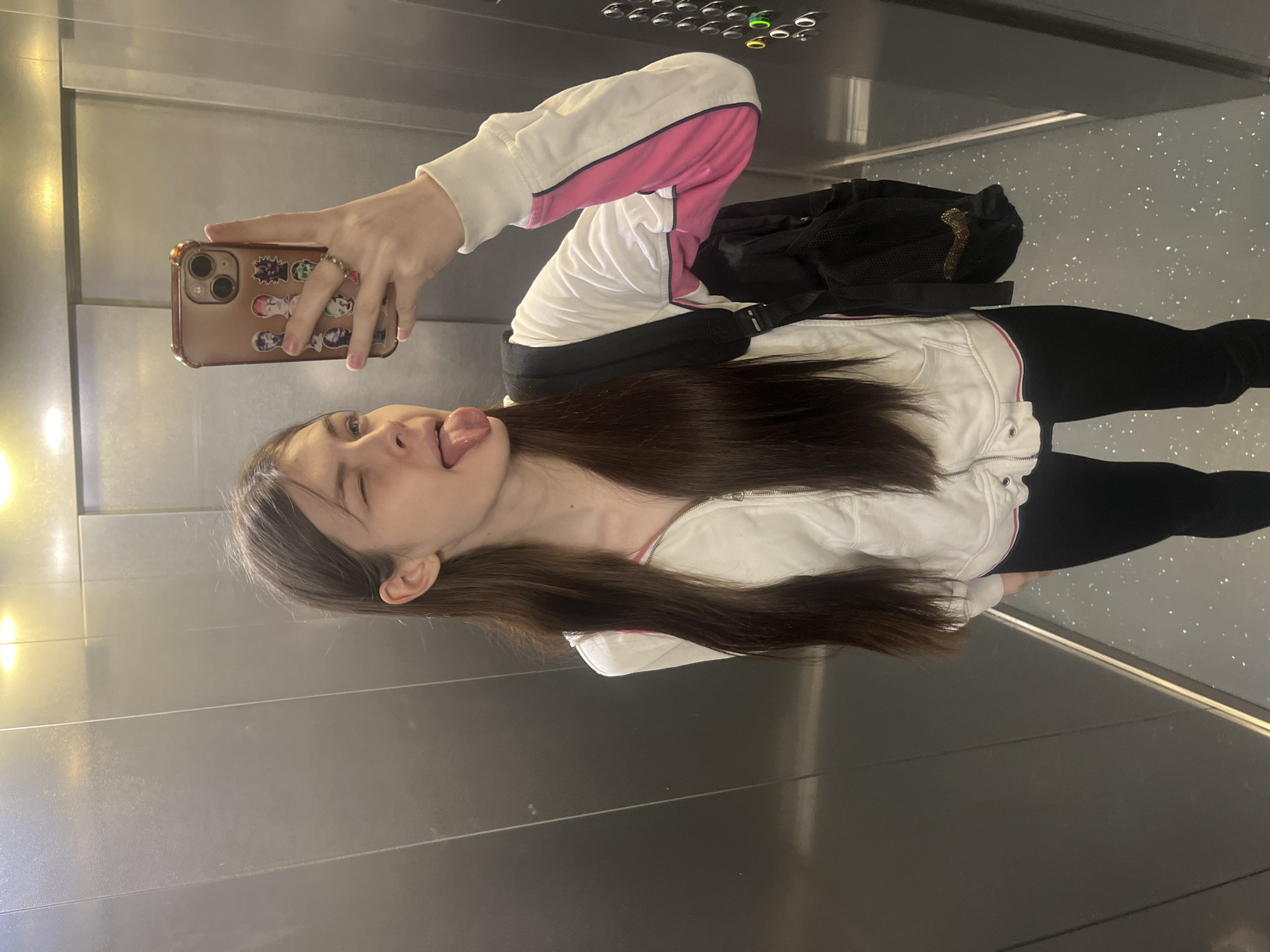 A young woman takes a selfie in an elevator, sticking out her tongue with a slight wink. She wears a white sweatshirt with pink accents and black leggings, and a black backpack is visible. She holds a phone with stickers on its case. The elevator has a control panel with lit buttons and is in a building in Saint Petersburg, Russia.