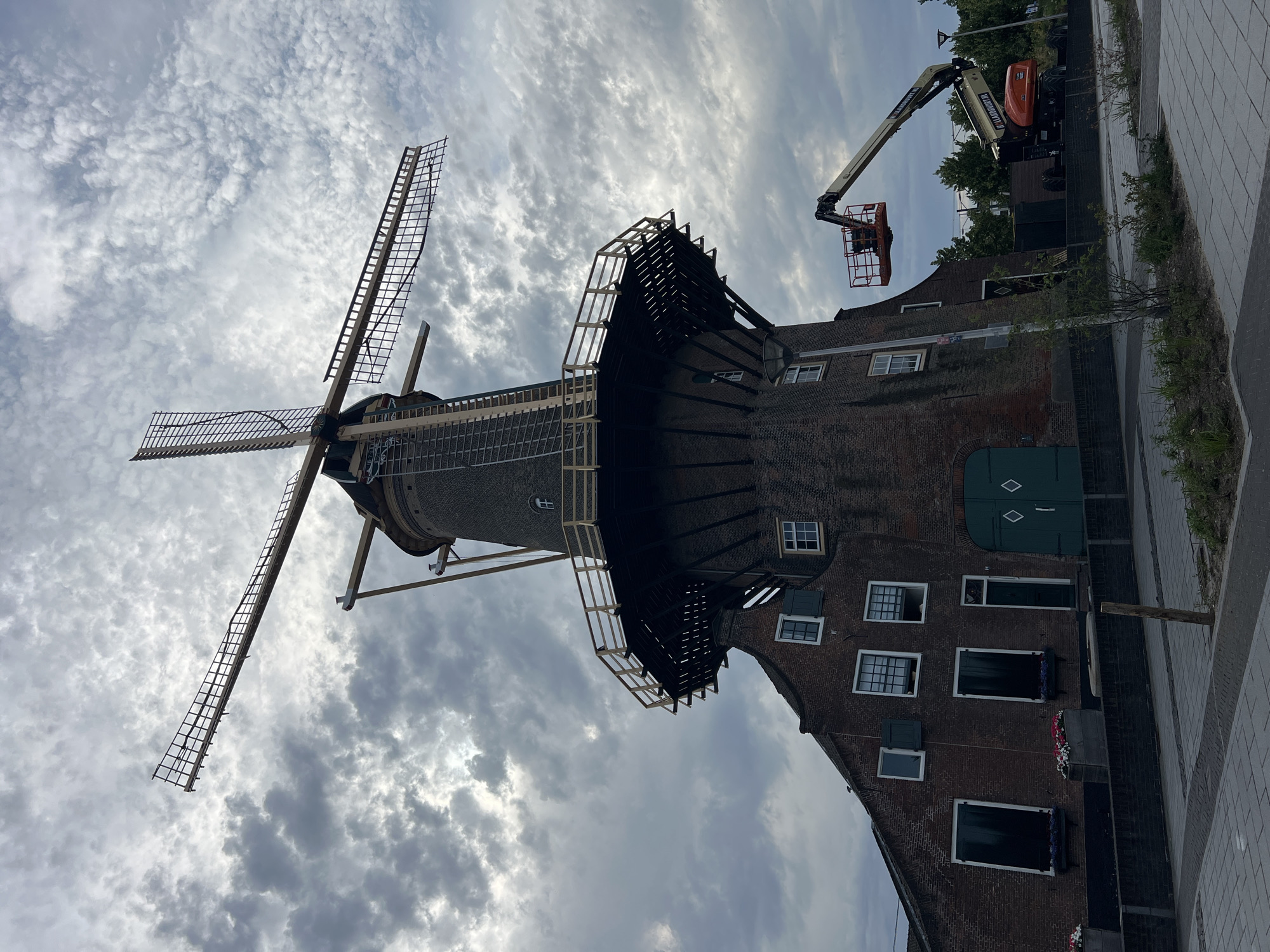 The image shows a traditional Dutch windmill undergoing maintenance. A cherry picker, a type of aerial work platform, is positioned next to the windmill, with its basket extended towards the upper part of the structure. The windmill itself is made of brick and has large wooden sails. The sky is cloudy, suggesting an overcast day. In the background, there are trees and a street. The overall scene appears to be in a developed area, possibly a town or city, given the paved walkway and the presence of a street lamp. The windmill seems to be a historical landmark or a functioning mill, with signs of recent activity indicating repairs or upkeep. The perspective is from below, looking upwards at the windmill and the sky. There is no visible text in the image.
