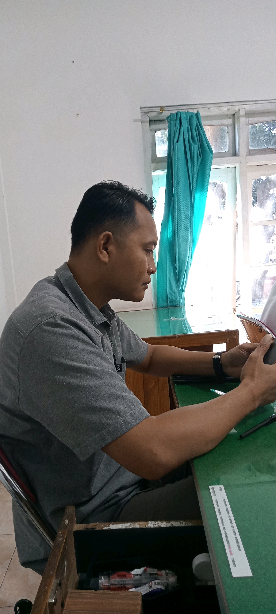 The image shows a man sitting at a green table in a room. He is wearing a grey collared shirt and appears to be looking at something in his hands, possibly a phone or tablet. Behind him, a window with teal curtains is visible, and the room's walls are white. The overall impression is of an indoor office or study space. There is no identifiable signage or context to definitively place the location within Begajah, Indonesia, though the style of the window and curtains might suggest a Southeast Asian setting.