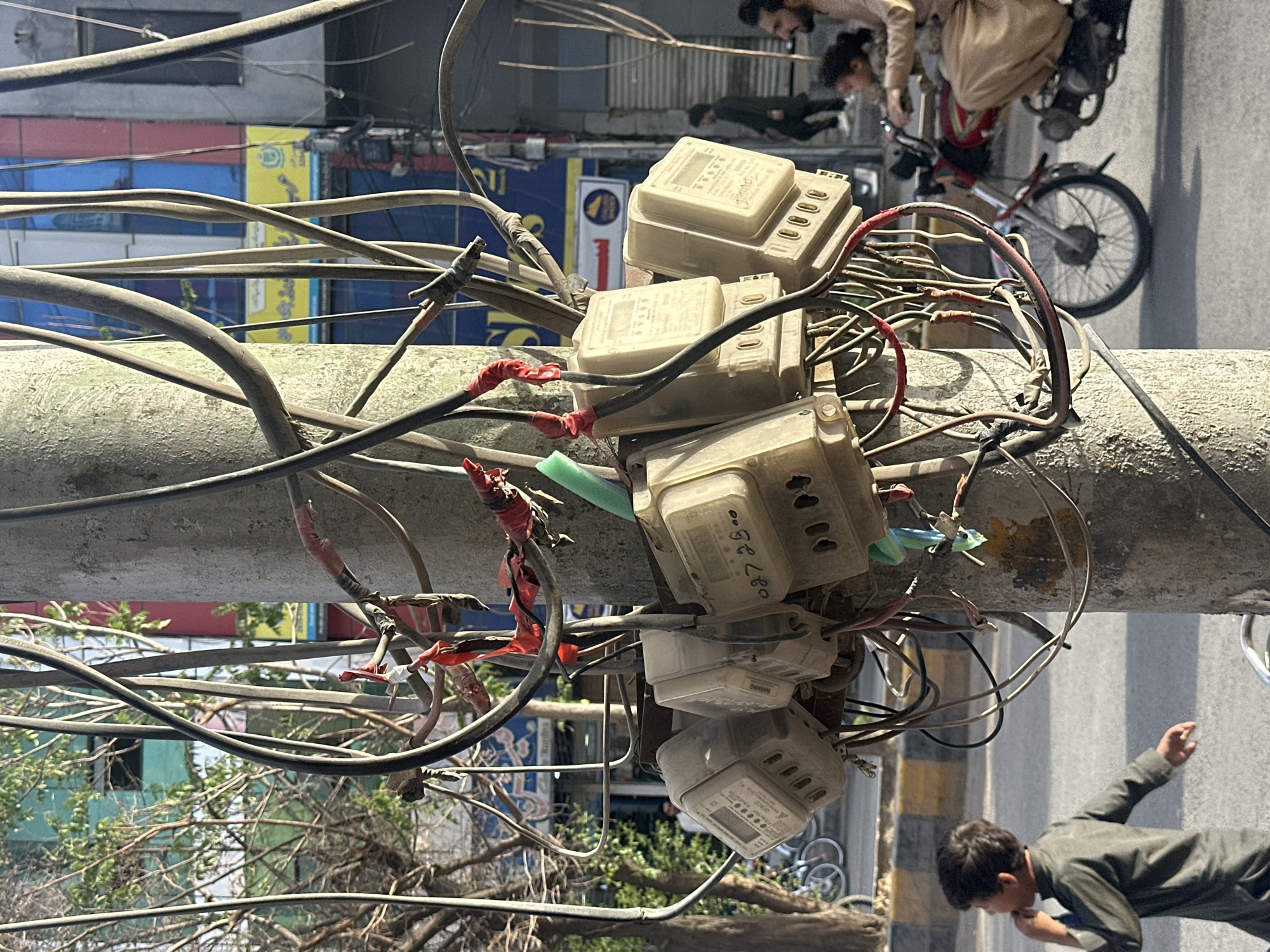 The image shows a utility pole in Quetta City Tehsil, Pakistan, laden with multiple electricity meters and a tangle of wires. The pole itself is concrete and weathered. Red electrical tape is used to secure some of the connections. Five electrical meters are stacked vertically on the pole. Several individuals are present in the background: two people are riding a motorbike, and a boy in a dark shirt is walking on the sidewalk below the pole. The environment includes buildings with colorful facades visible through the wires and tree branches. A bicycle is also visible in the background.