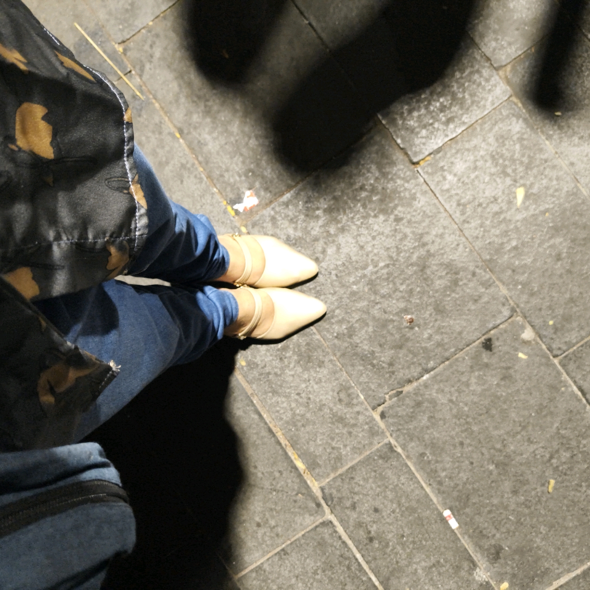 The image shows a person's legs and feet wearing blue denim jeans and light-colored pointed-toe heels with ankle straps. The person is standing on a gray tiled sidewalk. A dark patterned jacket is visible on the left side of the frame. Shadows suggest an outdoor environment, possibly at night or in a shaded area. The presence of discarded cigarette butts on the ground indicates a public outdoor space. This scene could be located in a street or public area within West Jakarta, Indonesia.