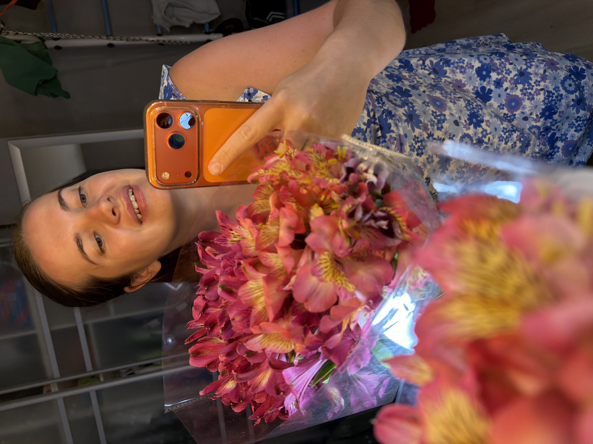 A young woman is depicted in a cheerful selfie, smiling broadly with her head tilted slightly, her blue eyes looking towards the camera. She has fair skin and dark hair. She is holding a smartphone in a bright orange case with her right hand, using it to capture the image.
Prominently displayed in the foreground and held by the woman is a large, vibrant bouquet of flowers, likely Alstroemeria (Peruvian lilies), featuring rich shades of pink, red, and yellow, with some purple accents. The flowers are encased in clear plastic wrapping, which shows some reflective glare.
The woman is wearing a light-colored dress adorned with a blue and purple floral pattern. The background is indoor, dimly lit, and largely out of focus, suggesting a casual home or indoor setting. There is no discernible text visible within the image. The overall impression is one of happiness and appreciation, possibly having just received the flowers. The context for this scene is Sochi, Russia.
