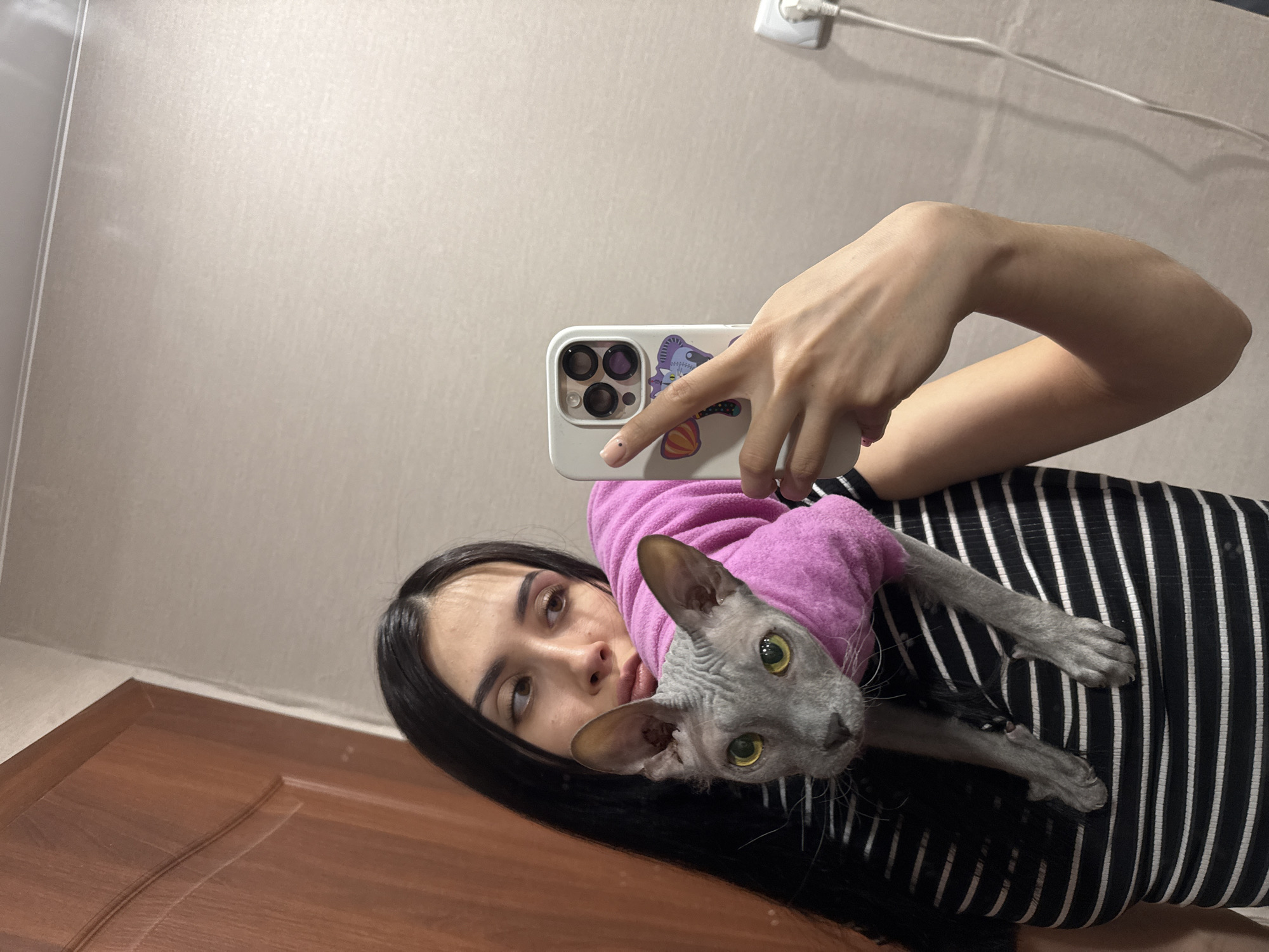 A fair-skinned person with dark hair is taking a selfie with a hairless cat. The cat is wearing a pink sweater. The person is wearing a striped black and white shirt. The person is holding a white cell phone with a butterfly sticker on it. The background is a beige wall with an electrical outlet. There is a brown door in the lower left corner. The location is indoors. The location is likely not in Votkinsk, Russia as there is no indication of this in the image.