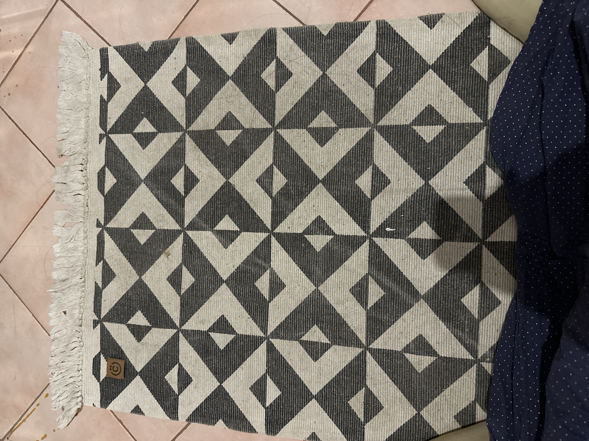 An overhead, full-frame shot shows a dark grey and off-white geometric patterned rug with a fringed edge. The rug is positioned on a tiled floor. In the upper left corner of the rug is a circular brown tag with a stylized "C" logo. Some light-colored fibers are scattered on the rug and the floor. The bottom of the image shows a dark blue fabric with a white polka dot pattern. The scene does not contain any identifying features or context to place it in Kalitino, Russia, or any specific city.