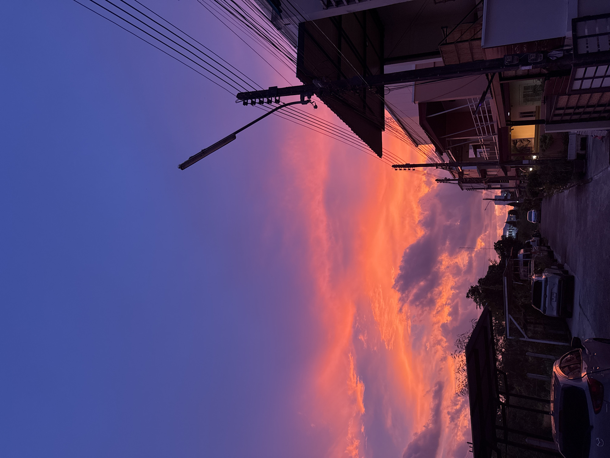 The image captures a street in Karon, Thailand, during a vibrant sunset. The sky is a gradient of deep blue at the top, transitioning into vivid shades of purple and fiery orange and pink where the sun is setting. Clouds are illuminated by the sunset, adding texture and depth to the sky.
On the right side of the street, a multi-story building is visible, with balconies and windows. A utility pole stands prominently, adorned with numerous electrical wires and a street lamp. Several other utility poles and wires are scattered throughout the scene, indicating the presence of infrastructure.
The street itself is paved, and several parked cars are lined up along the side. One car, a silver hatchback, is in the foreground on the left, its silhouette dark against the bright sky. Further down the street, other vehicles, including what appear to be SUVs and vans, are parked. On the left side of the street, there are covered carports and some vegetation, including trees. The overall impression is that of a residential or mixed-use urban environment bathed in the dramatic colors of a tropical sunset. There are no people visible in the image.
