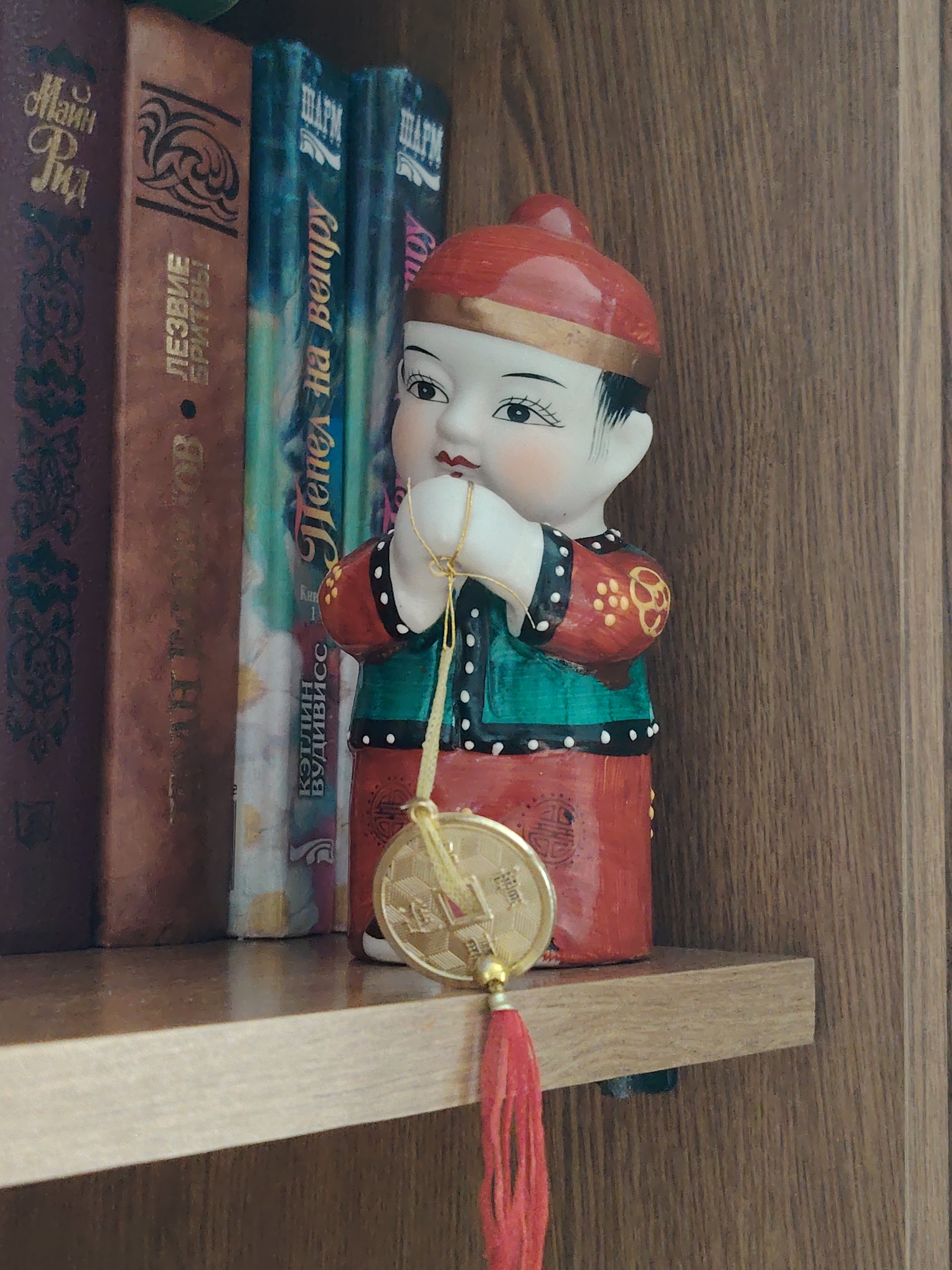 A ceramic figurine of a child in traditional East Asian attire stands on a wooden shelf next to several books. The figurine wears a red cap with gold trim, a green vest over a red garment adorned with gold and white polka dots, and red trousers. It holds a gold-colored coin with intricate designs and a red tassel hanging from it. The books are arranged vertically, with their spines facing outwards. The spines feature text and decorative elements. The background is a wooden surface with a visible grain pattern. The scene appears to be an indoor setting, possibly a bookshelf in a home or library, in Rostov-on-Don, Russia.