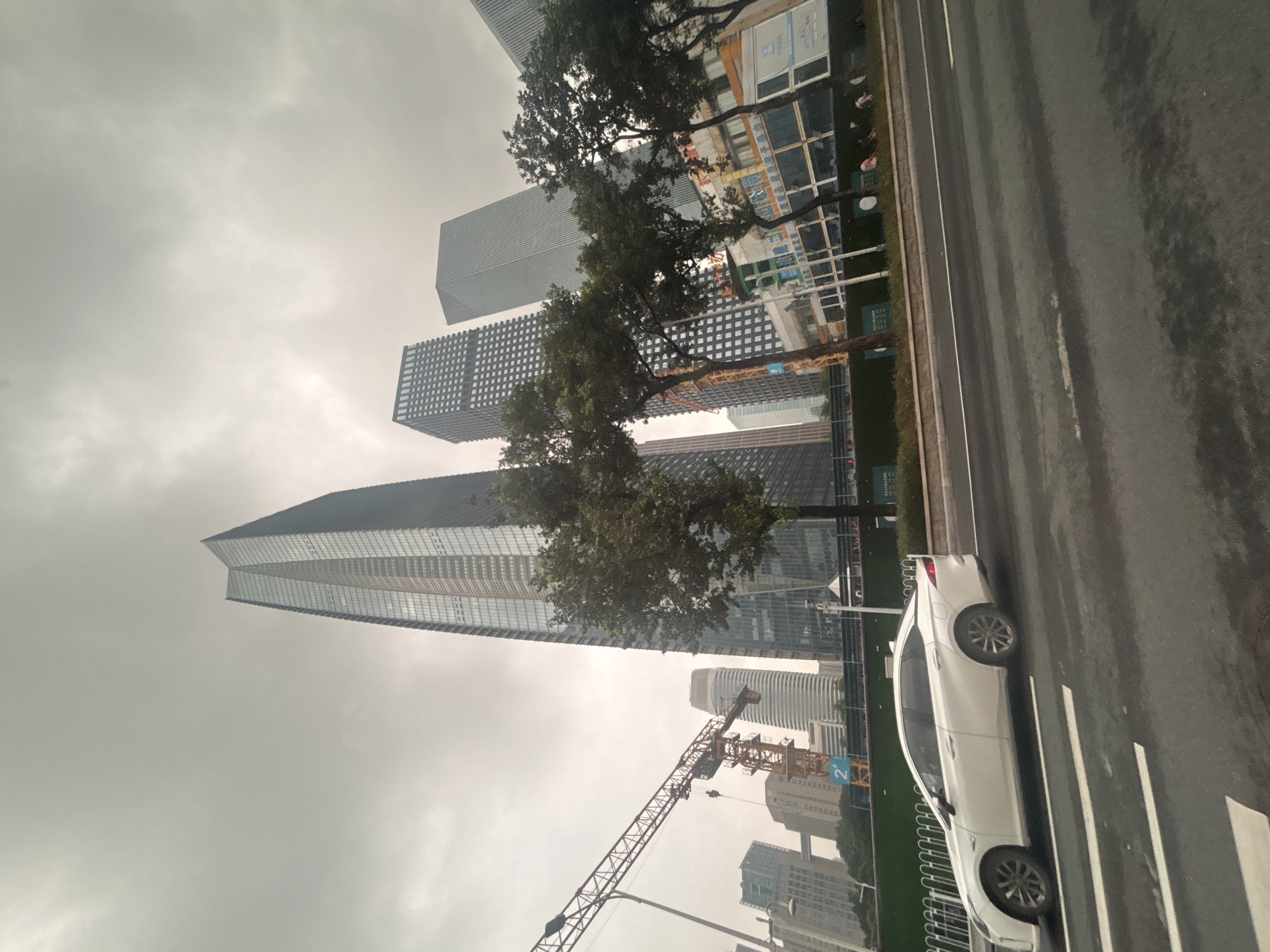 A wide-angle, eye-level shot captures a street scene in Tianhe District, China, on an overcast day. Dominating the background are several modern skyscrapers, characterized by glass facades and geometric designs. The tallest, a sleek, tapered building, stands prominently on the left. To its right are other tall, rectangular structures with patterned window grids and smooth, angled surfaces. A large construction crane, orange and yellow, is visible in the mid-ground, suggesting ongoing development. Trees with dense green foliage line the area between the road and the buildings. In the foreground, a white sedan is traveling on the right side of a multi-lane road. The road surface shows faint white lane markings and appears wet. A low green fence separates the road from a grassy area with several small figures of people scattered throughout, some appearing to be seated. The sky is uniformly gray and cloudy.