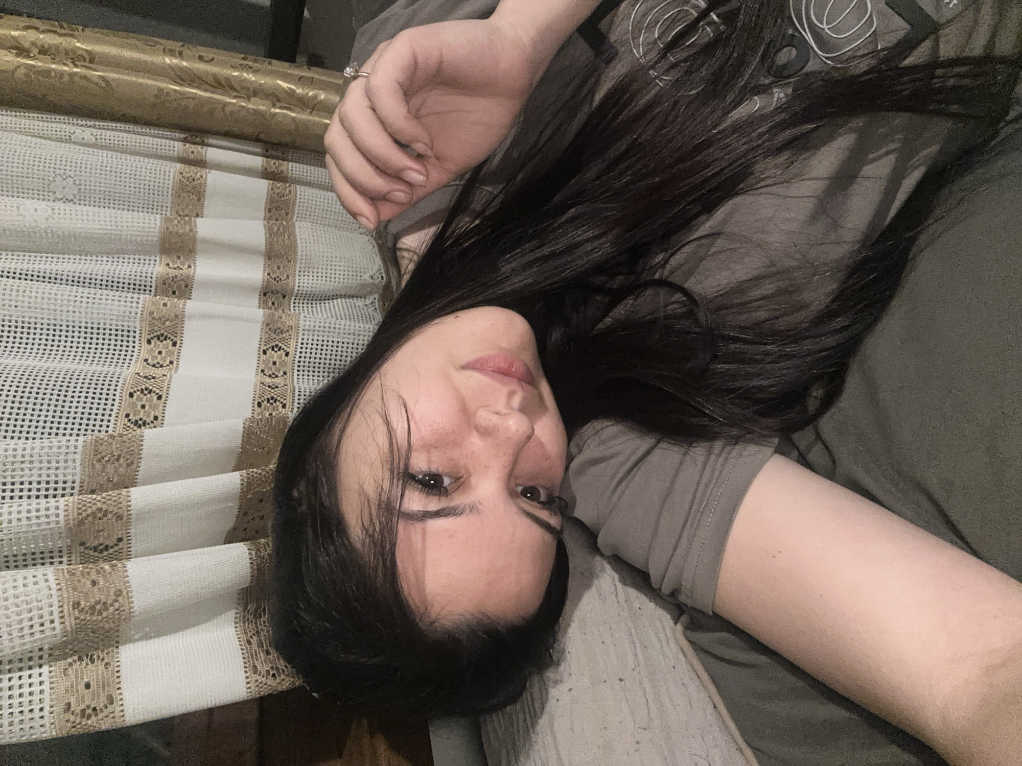 The media file presents an indoor portrait of a young woman, likely in Chubra, Bulgaria, lying down and looking directly at the camera. She has long, dark hair that spreads around her, fair skin with noticeable freckles across her nose and cheeks, and dark eyes. She is wearing a dark grey or olive green short-sleeved t-shirt, which has faint white abstract or circular patterns visible on the upper left side.
Her right arm is bent with her hand resting near her head, showcasing a ring on her ring finger. Her left arm is partially visible, resting by her side. She is lying on soft, dark grey bedding or cushions.
To her left, a decorative, off-white fabric is prominently displayed, possibly a curtain or bed runner. This fabric features a grid-like mesh pattern interspersed with distinct horizontal bands of intricate golden-brown lace-like design. Behind this patterned fabric, a rolled-up object with a shiny golden, ornate pattern is visible, suggesting a decorative element like wallpaper or another fabric roll.
The lighting is soft and appears to be artificial, typical of an indoor setting, possibly evening or night. Her expression is calm and direct, engaging with the viewer. There is no discernible text visible in the image.