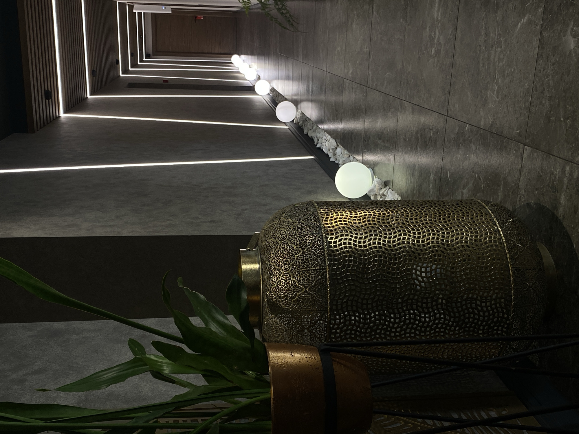 The image displays a modern, dimly lit interior hallway within a building in Votkinsk, Russia. In the left foreground, a green potted plant in a bronze planter on a dark stand is positioned beside a large, ornate golden-bronze cylindrical lantern featuring intricate latticework patterns. The hallway floor is covered in dark grey rectangular tiles. The walls are textured grey, each featuring embedded vertical strip lighting that extends into the distance, converging with the perspective. The ceiling, partially visible further down the corridor, incorporates wooden slats and recessed linear lighting. Along the left side of the hallway floor, a series of at least seven spherical white luminous orbs are arranged on a bed of white decorative pebbles. Multiple wooden doors are visible on the right wall in the background, with one door in the far distance exhibiting a red fire extinguisher mounted next to it. No individuals are present.