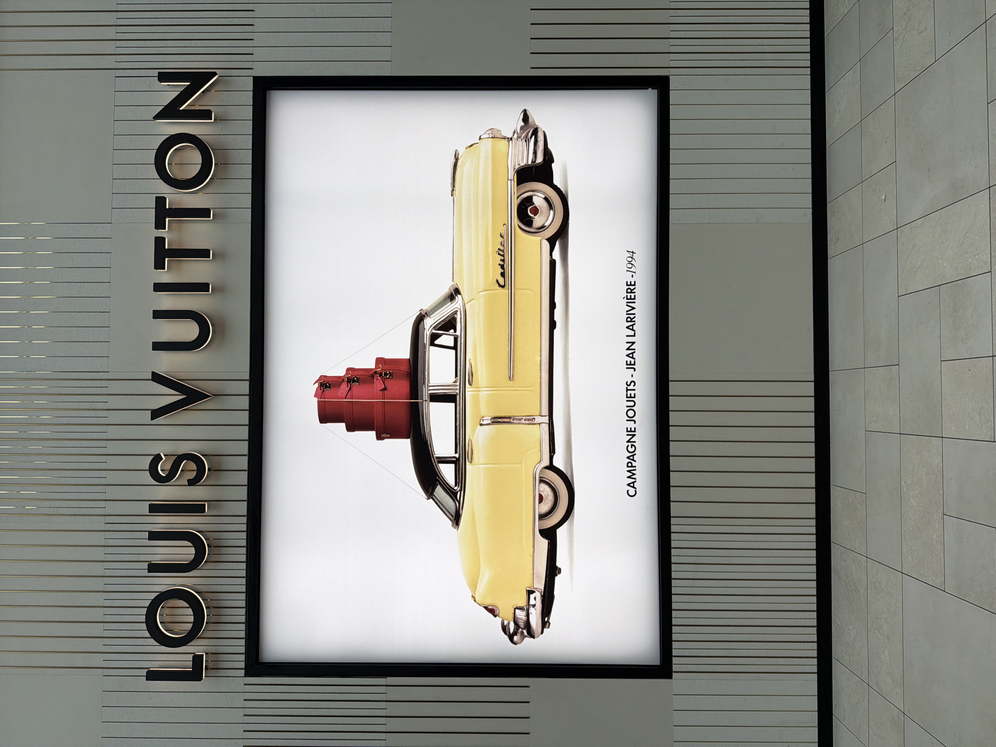 The image depicts an advertisement for Louis Vuitton in the city of Mount Royal, Canada. The Louis Vuitton sign, with large illuminated letters, is attached to a wall with horizontal grey and beige stripes. Adjacent to the sign is a framed advertisement featuring a vintage yellow car, possibly a Cadillac, with stacked red Louis Vuitton trunks tied to the roof. The image inside the frame also has text "CAMPAGNE JOUETS - JEAN LARIVIERE - 1994". The wall to the right of the advertisement is composed of large, light-colored rectangular tiles.