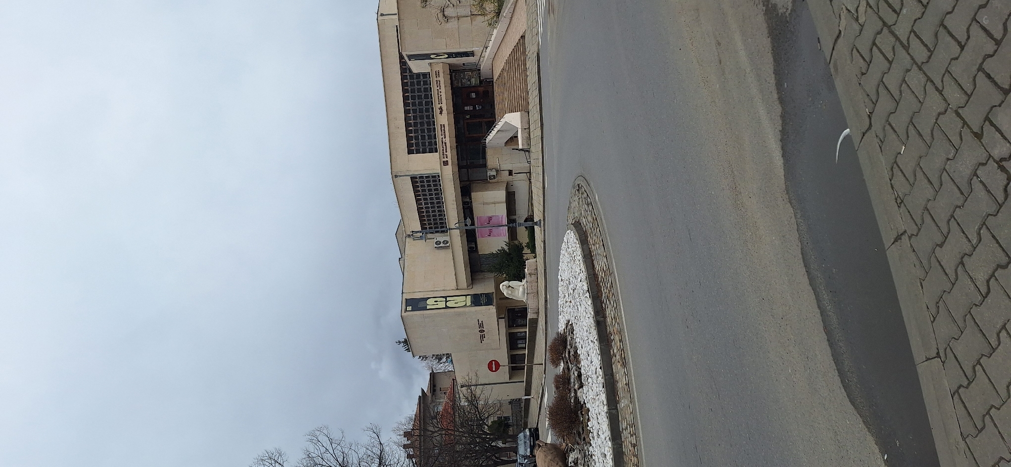 The image shows a street scene in Kazanlak, Bulgaria. A large, multi-story building dominates the left side of the frame. It has a light-colored facade and a grid of windows. A small roundabout with white stones and sparse shrubbery sits in the road in front of the building. The road itself is asphalt. The right side of the frame is dominated by a paved area of gray bricks. The sky is overcast. There are bare trees in the lower left corner of the image. A "no entry" sign is visible on the building near the street level.