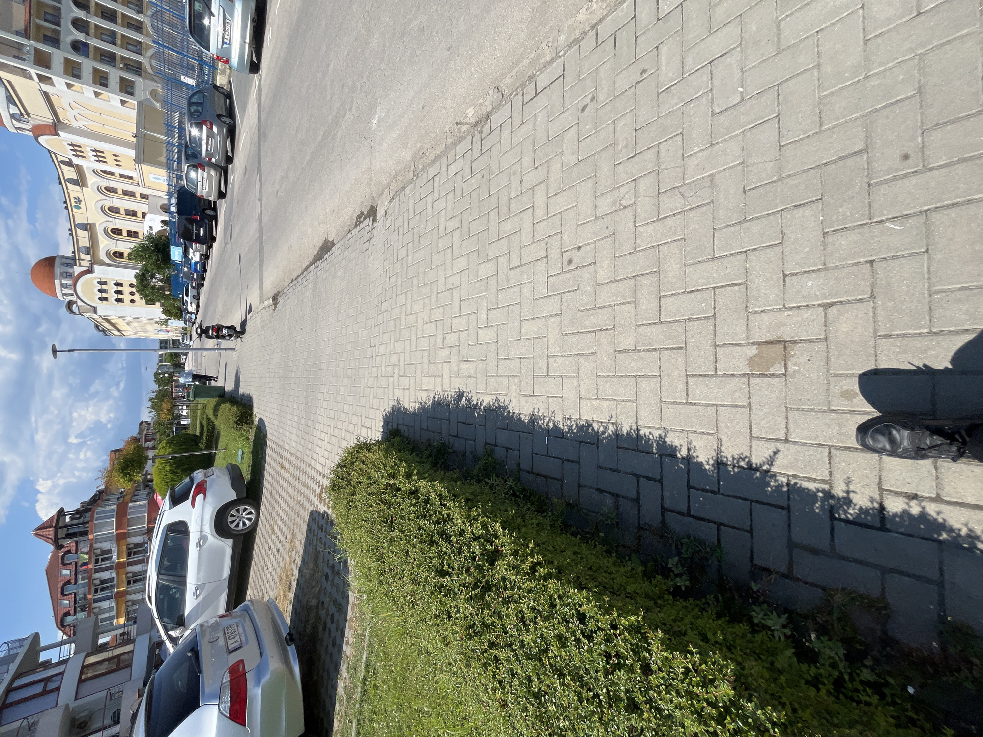 The image captures a street scene in Obzor, Bulgaria, on a sunny day with scattered clouds. The viewpoint is from ground level, looking down a paved sidewalk towards a street lined with parked cars.
On the left side of the image, a neatly trimmed green hedge runs alongside the sidewalk. Behind the hedge, a silver car is parked. Further down the street, a white SUV is parked next to a building with a distinctive red, multi-tiered roof. Adjacent to the SUV is a modern building with a beige facade, featuring numerous arched windows and balconies, topped with a large, orange-domed structure.
The street itself is paved with asphalt and has several cars parked along its right side. The parked vehicles include a dark blue minivan, silver hatchbacks, and a silver sedan closer to the foreground. A black scooter is parked on the sidewalk near the curb, with a person in a blue shirt walking away from it further down the street.
The sidewalk is constructed of light gray rectangular paving stones laid in a herringbone pattern. The paving stones are slightly uneven in places, with some darker stains and a small piece of white debris visible. The shadow of the person taking the photo, along with their black shoes, is cast on the sidewalk in the foreground. The overall impression is of a well-maintained, urban environment in a sunny climate.