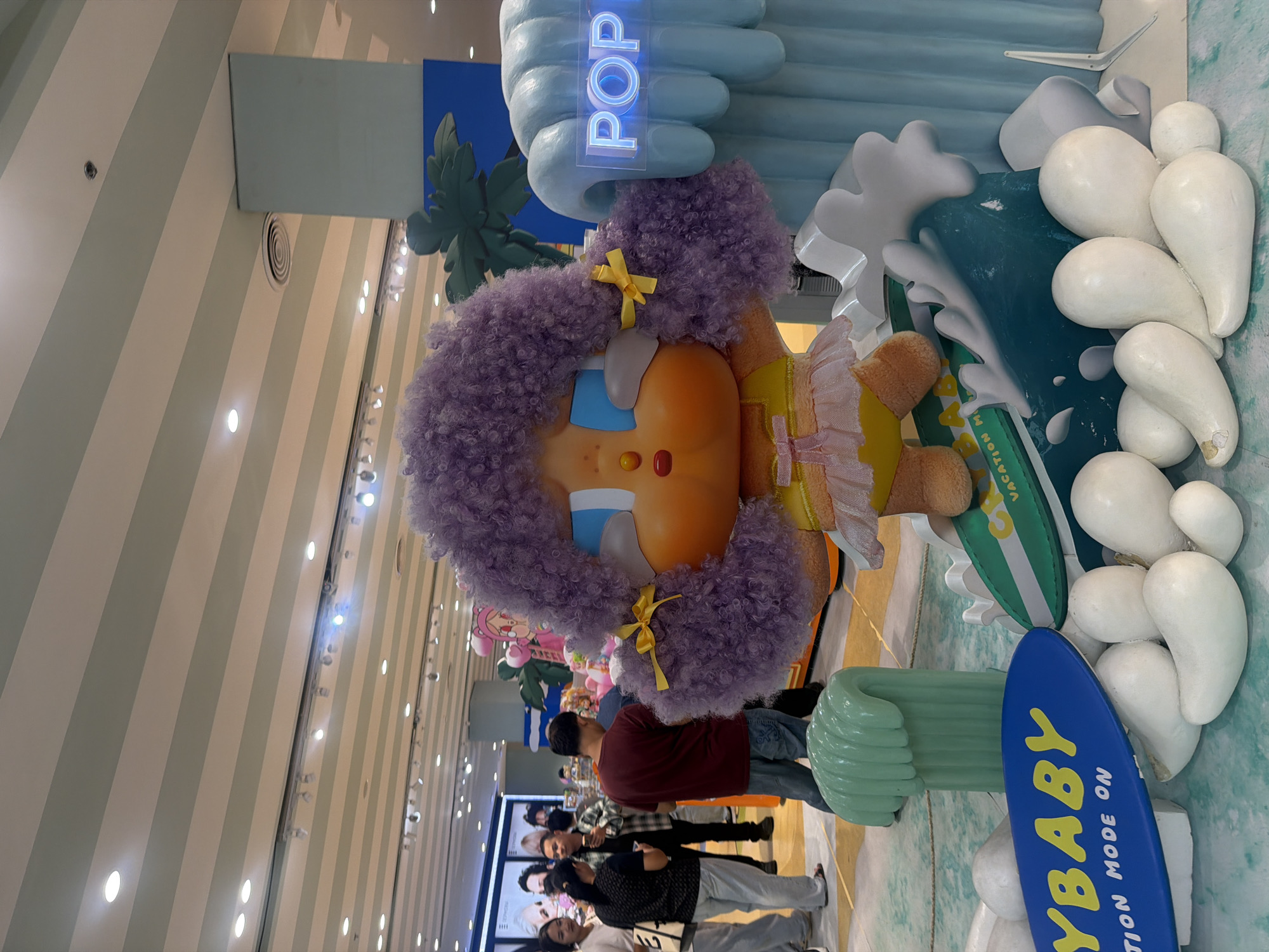 An indoor scene with a large display of a cartoon character with purple curly hair, big blue eyes, and an orange face, wearing a yellow swimsuit with a pink ruffled skirt. The character is standing on a surfboard that is positioned on a blue wave sculpture. In the background, there are people browsing in what appears to be a retail store. The display also features white, rounded objects resembling pebbles or clouds, a green tubular structure, and a blue sign with white text that reads "POP". Another blue surfboard with yellow lettering that says "YBABY" is in the foreground. The ceiling has a striped pattern of light green and white with recessed lighting. The overall environment suggests a shopping mall or a retail exhibition space in Wichit, Thailand.