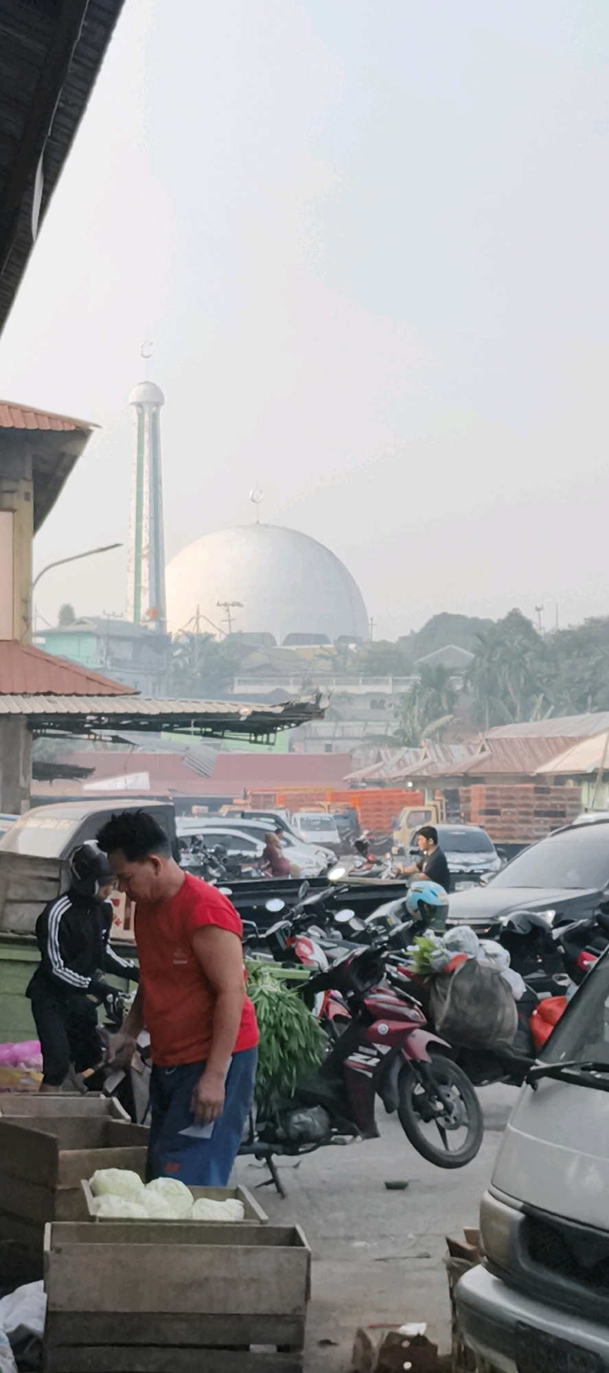 The image depicts an outdoor market scene in Jambi City, Indonesia. In the background, a prominent mosque with a large dome and a tall minaret is visible, partially obscured by haze. The foreground is a bustling marketplace with several individuals engaged in various activities.
To the left, a person wearing a black tracksuit with white stripes is interacting with another individual in a red t-shirt and blue pants. The person in the red shirt is looking down, holding a paper. In front of them, wooden crates filled with what appear to be cabbages are displayed.
Numerous motorcycles are parked and in motion throughout the scene, indicating transportation and activity. Some motorcycles are laden with goods, suggesting commercial purposes. Several cars and trucks are also present, some parked and others appearing to be moving through the market area.
The environment suggests a daytime setting, possibly morning or late afternoon, due to the soft lighting and the haze in the atmosphere. The architecture of the market stalls, with their corrugated metal roofs, is typical of a local market in the region. The overall impression is one of a busy, active urban market in Jambi City.