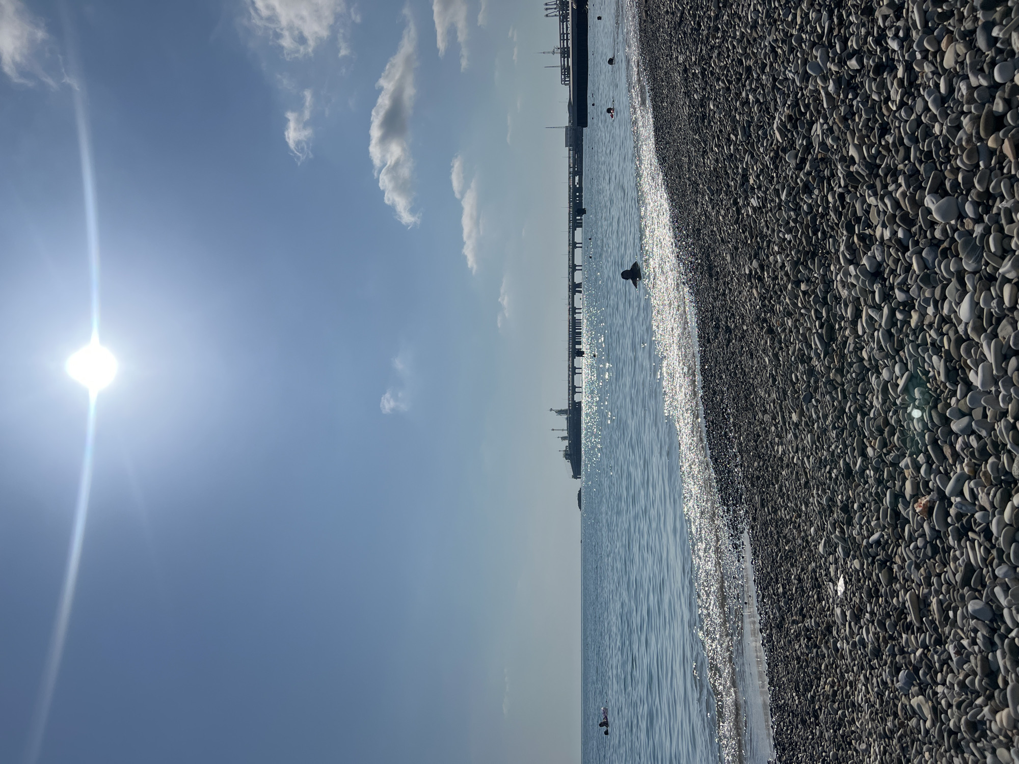 The image depicts a sunny day at a pebble beach in Kroyanskoye, Russia. The sun is bright and high in the sky, creating a glare on the water. The sea is calm and clear, with small waves gently lapping at the shore. In the distance, a long pier extends into the sea, with structures that appear to be part of an industrial facility. Several people are visible in the water, swimming and enjoying the beach. The foreground of the image is dominated by a dense scattering of grey and white pebbles, typical of the coastline in Kroyanskoye. The overall atmosphere is serene and suggests a leisurely summer day.