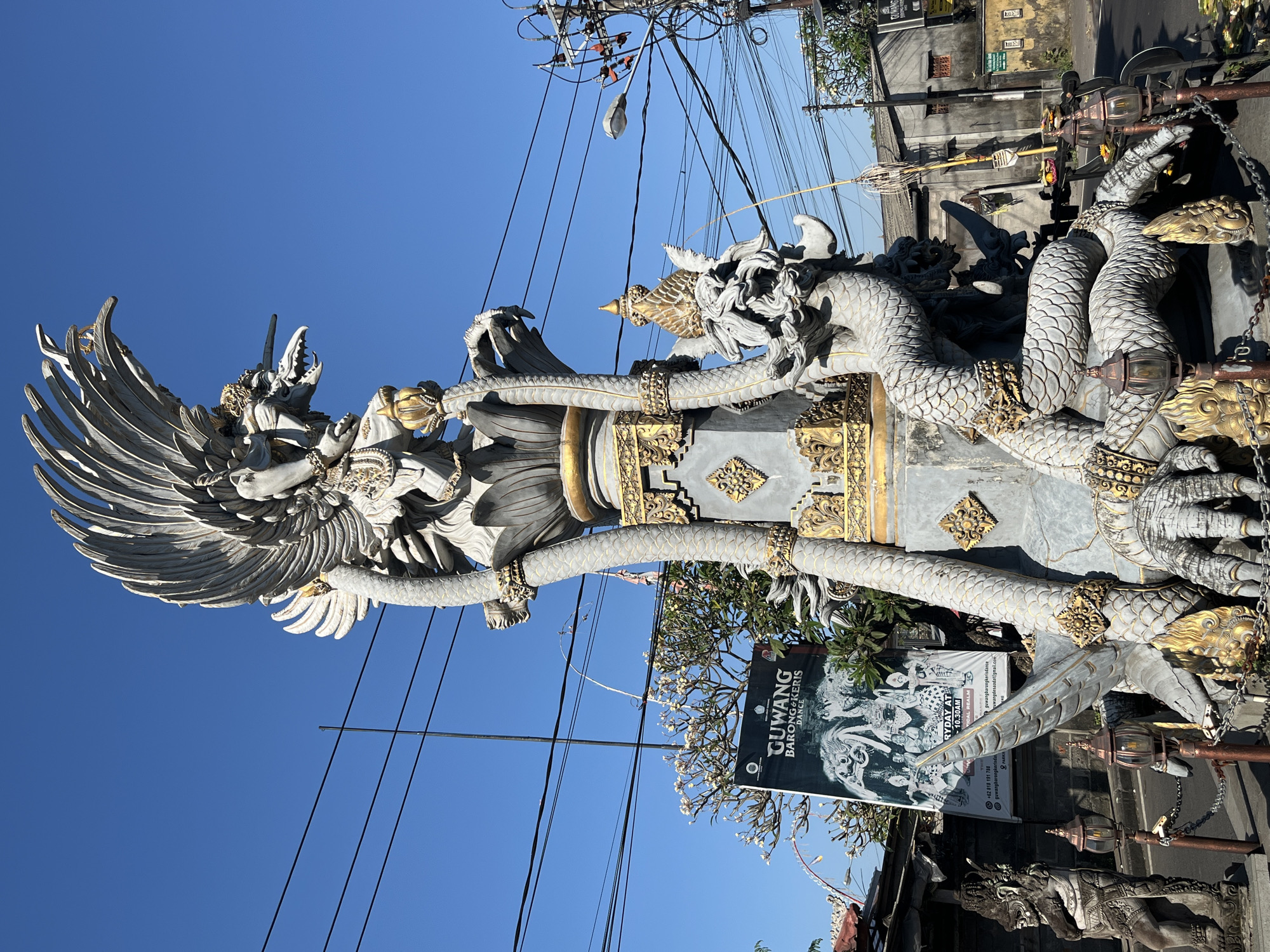 An outdoor, eye-level, medium shot shows a detailed, grey stone statue of a mythological Garuda bird with large feathered wings and a dragon entwined around it. The statue is ornately decorated with gold accents on the base, dragon scales, and bird's headdress. It stands on a pedestal with a lotus flower design at the top. The statue is located in the city of Sukawati, Indonesia, a fact indicated by a poster in the background advertising "GUWANG BARONG & KERIS DANCE" with a phone number and website. The background features a clear blue sky, power lines, a street light, buildings, trees, and another stone statue to the left of the main statue. The statue is surrounded by a decorative chain and several lanterns. The sun is shining brightly, casting shadows on the scene.
