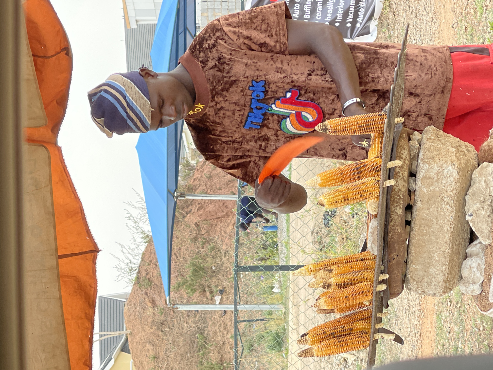 An outdoor scene in Abuja, Nigeria, features a man roasting corn on the cob. The man, appearing to be in his 20s or 30s, wears a brown, velvet-textured short-sleeved shirt with a prominent, colorful "TikTok" logo embroidered on the front. He also wears a striped cap in shades of blue, beige, and brown, and red shorts.
He is actively engaged in his task, holding an orange spatula or flipper to turn multiple ears of corn. The corn is arranged on a rustic grilling setup, consisting of a wooden or metal frame supported by large stones, suggesting an open-fire method. Many ears of corn, some showing charring, are visible on the grill.
The setting is bright daylight, with a clear sky. Two tents, one orange and one blue, provide shade. A chain-link fence separates the grilling area from a background of dry, sparsely vegetated ground and a small hill. Distant buildings are also visible. On a white banner in the upper right background, fragmented text advertising services like "Auto c", "Buffing", "ceram", "headlig", "Interior", "Vacuum", and "Auto dia" can be seen, indicating a nearby commercial establishment, possibly an auto detailing business. The overall scene depicts a common roadside or market food vendor.