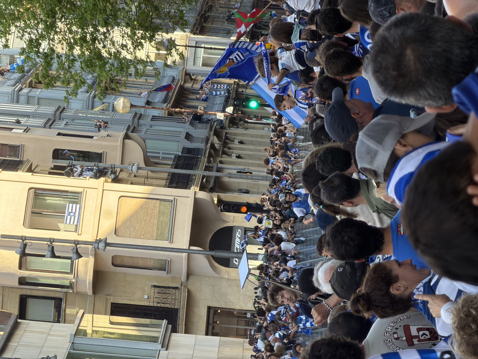 This image depicts a lively street scene in San Sebastián, Spain, during what appears to be a public celebration or parade. A large crowd of people fills the street, with many individuals wearing blue and white striped clothing, indicating support for a local sports team, most likely Real Sociedad. Numerous flags are visible, including several blue and white banners, some with a white crest, and the red, green, and white Basque flag (Ikurriña). A red, blue, and yellow flag, possibly Venezuelan, is also seen on a balcony.
The setting features multi-story buildings lining the street, with spectators observing from balconies. The architecture is typical of a European city. The lighting suggests late afternoon or early evening, with golden sunlight illuminating the scene and casting soft shadows. Many people in the crowd are holding up mobile phones, capturing photos or videos of the event.
A traffic light displays a green signal, while another shows orange. Part of a building facade features the text "DIMAS" or "CIMAS." On a t-shirt worn by a person in the crowd, the phrase "ALWAYS FRESH!" is visible alongside a graphic. The overall mood is one of excitement, community, and celebration.