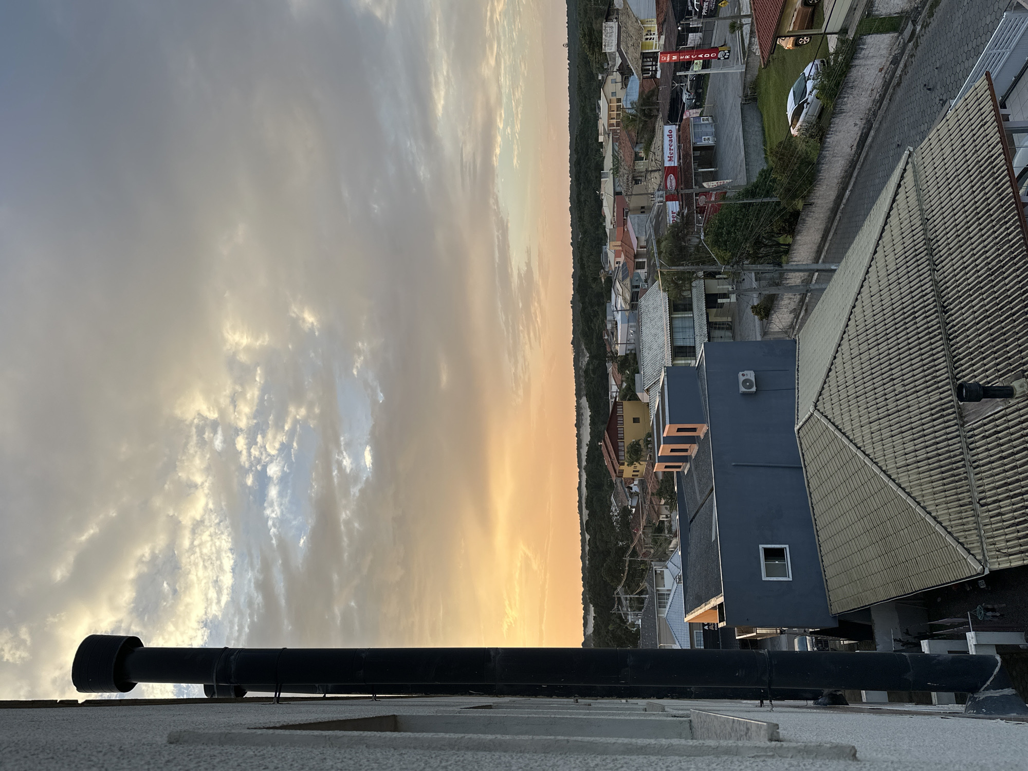 This image captures a sunset over a coastal town in Florianópolis, Brazil. The sky is filled with dramatic clouds tinged with orange and yellow hues. In the foreground, a large black chimney pipe runs vertically along the side of a textured building. Below, a cluster of residential and commercial buildings with diverse rooflines and colors stretch towards a dense line of dark green trees. In the mid-ground, a street is visible, paved with cobblestones, with a white car parked on the side and a few birds on the ground. Signs for "Mercado" indicate a commercial area. The overall scene depicts a residential and commercial area at dusk.