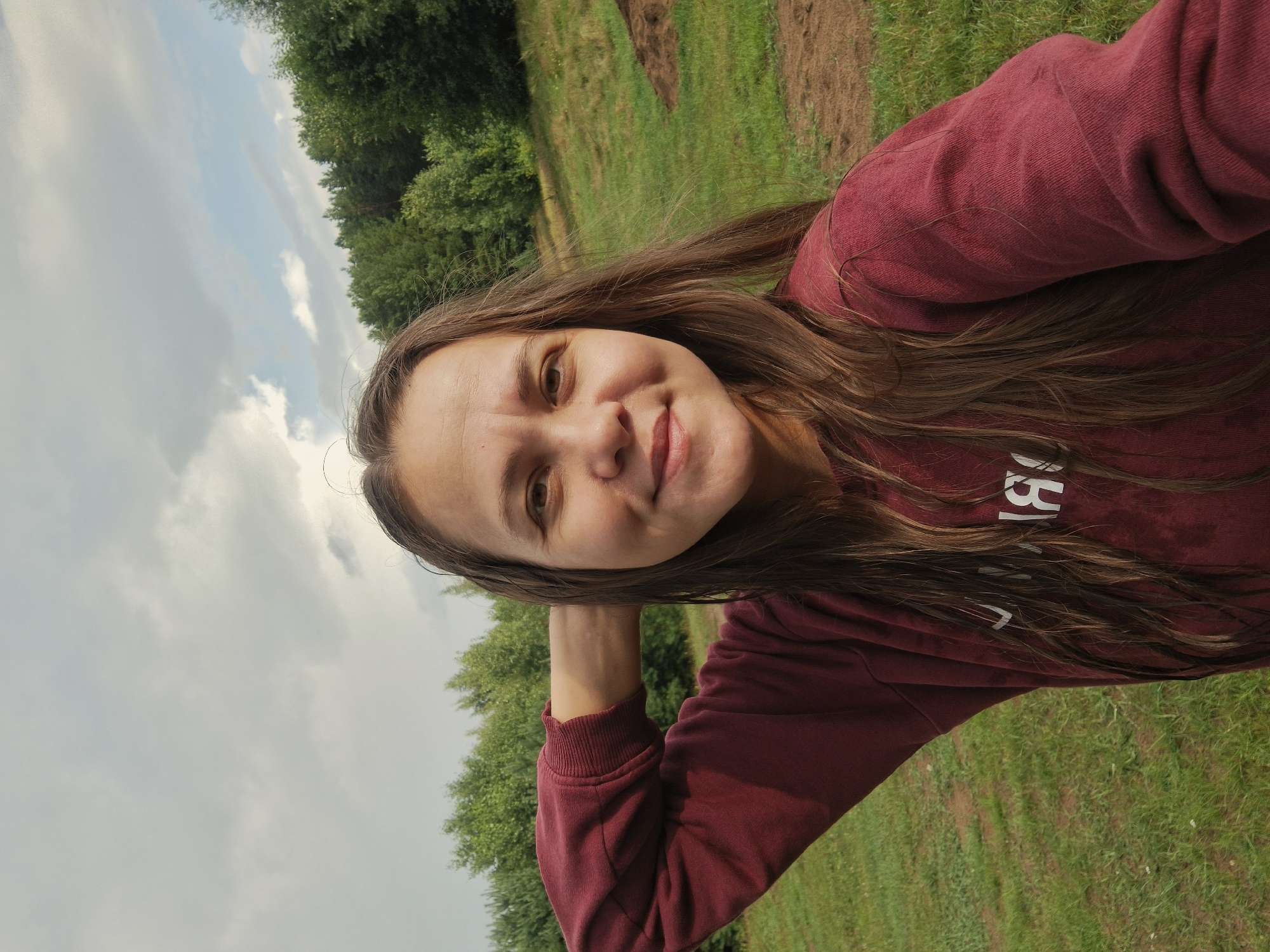 The image shows a medium shot of a woman with long, brown hair, taking a selfie outdoors. She is wearing a maroon sweatshirt and has her left arm raised, with her hand behind her head. The background consists of a grassy field with trees and a cloudy sky. The location is identified as Plishkino, Russia. There is a small pile of dirt in the middle ground. The woman is smiling gently at the camera.