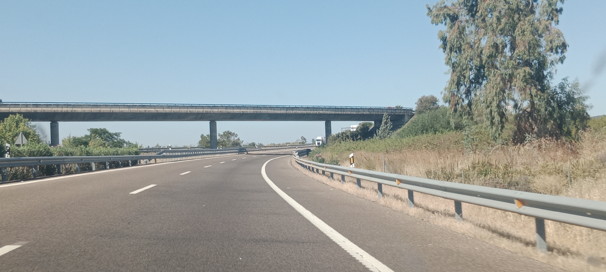 The image shows a highway passing under an overpass. The highway has two lanes in the visible direction of travel, marked with white lines. A metal guardrail runs along the right side of the highway, separating it from a grassy embankment with a large tree. The sky is clear and blue. The overpass appears to be made of concrete and is supported by pillars. In the distance, there are some trees and vegetation. A white truck is visible on the overpass. There are no people visible in the image. The scene is consistent with a rural highway in Spain, potentially near Torrefresneda.