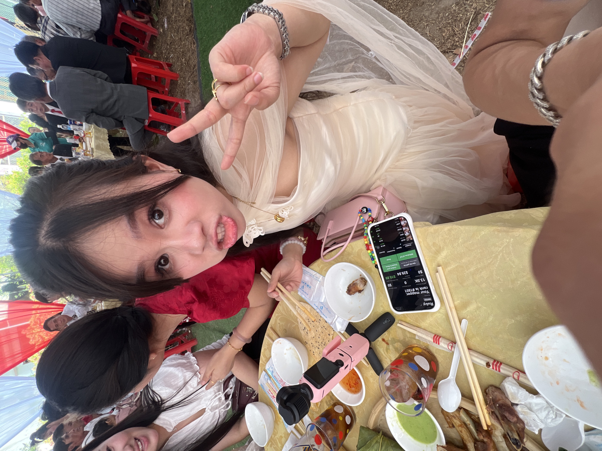 An overhead selfie captures a young woman at an outdoor gathering in Tay Son Commune, Vietnam. She is positioned at the top of the frame, lying on her side or leaning over a table, wearing a light-colored, possibly off-white, formal dress with sheer fabric. She makes a peace sign with her right hand and looks playfully towards the camera.
The scene below her shows a lively outdoor event with multiple people seated at tables covered with yellow tablecloths, likely enjoying a meal. Many attendees are dressed in suits or semi-formal attire, while others wear more casual clothes, including red and white traditional-looking garments. Red, white, and blue fabric decorations are visible in the background, suggesting a festive occasion. The ground appears to be covered in artificial grass.
On the table in front of the main subject, various items are scattered: empty and partially eaten bowls, chopsticks, a pink portable camera or mini tripod, glasses, and saucers with dips. A white smartphone is prominently displayed, showing an app interface. Visible text on the phone screen includes "Ruby," "MAPPER," "Your mapper, rank is #1801," "13.5K," "$0," "$29.84 $10.89," "Get 29 PATTN," and "Withdraw ATTIK." The bright lighting suggests it is daytime. The overall atmosphere appears social and celebratory.
