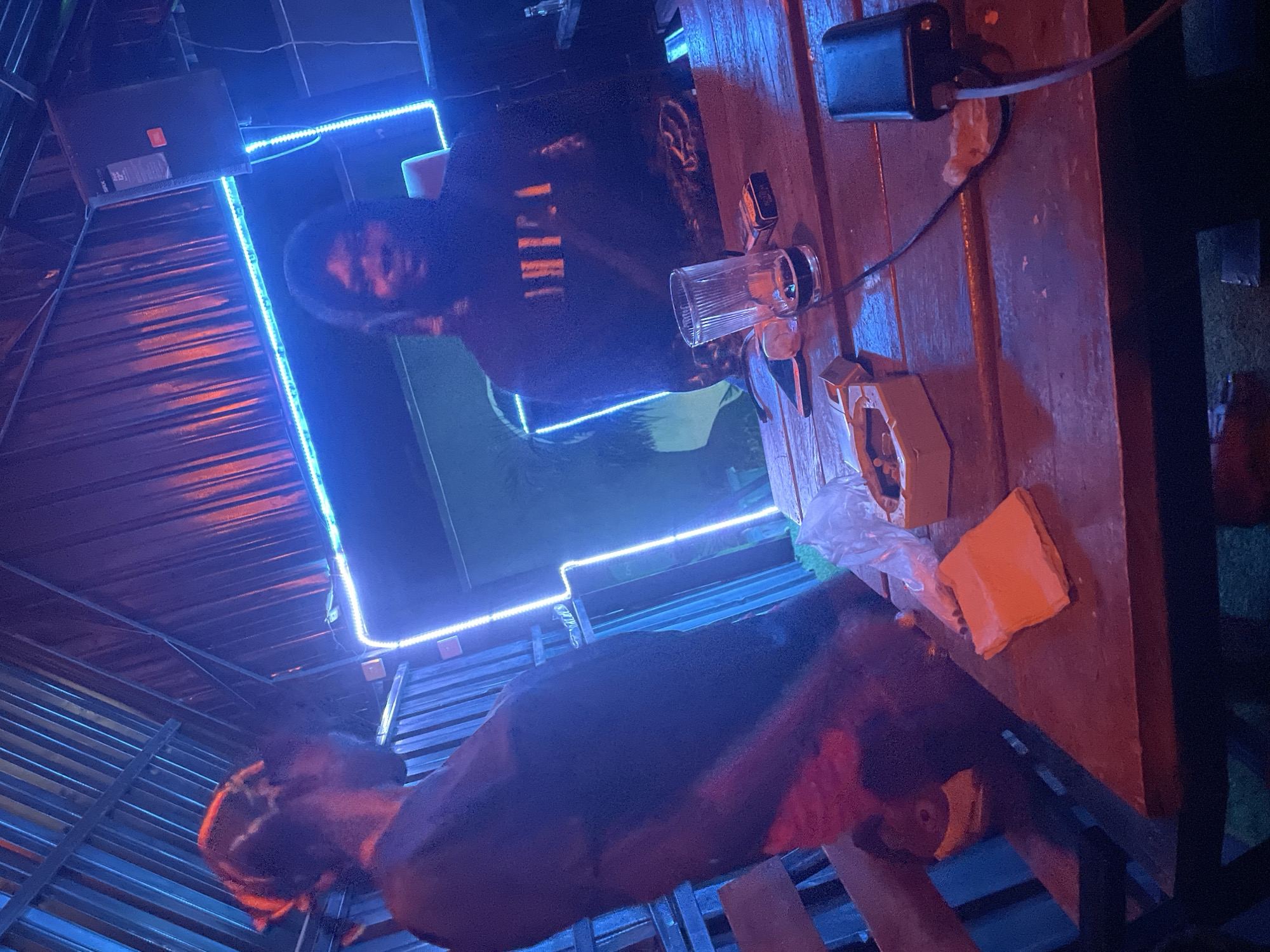 The image shows an indoor setting in Gura Top, Nigeria, possibly a bar or restaurant. Two individuals are visible, although their features are obscured by the lighting. One person is standing in front of a square frame illuminated with blue LED lights. Another person is positioned below, partially obscured. A wooden table is present, bearing a glass, a box, a black object with a wire, and what appears to be an orange napkin or cloth. The room has a ceiling composed of metal or corrugated sheets. The lighting is a mix of blue LED strips and warmer, redder tones.