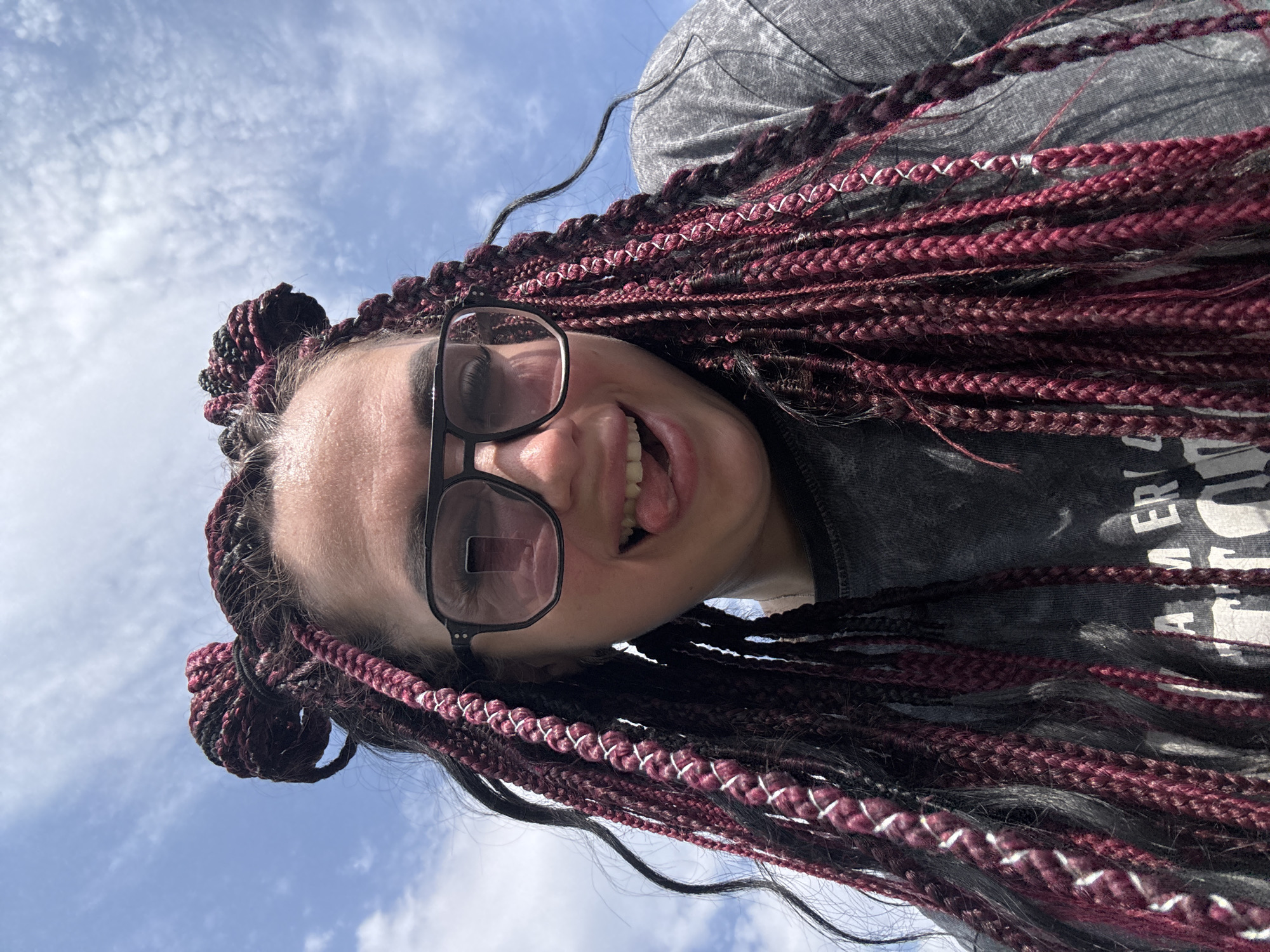 A young woman with burgundy and black box braids styled in two high buns is shown in a close-up, upward-angled selfie against a bright blue sky with scattered white clouds. She is wearing large, dark-rimmed glasses with pinkish lenses and a dark grey t-shirt. Her mouth is open, and her tongue is sticking out playfully. The image appears to be taken outdoors. There is no visible indication of the city of Tolyatti, Russia.