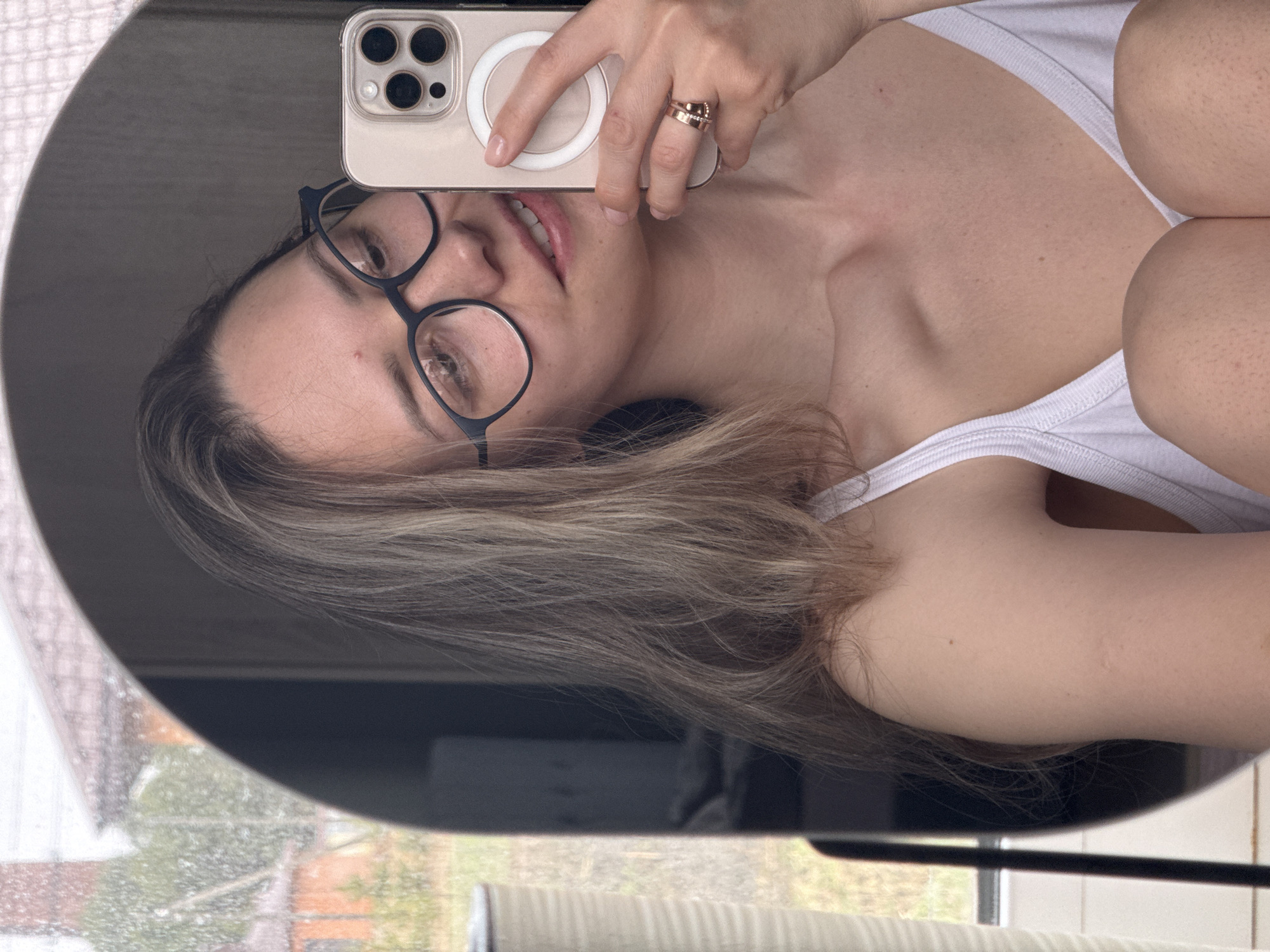 A woman is taking a selfie in a round mirror. She is wearing glasses and a white tank top, and is holding an iPhone. The background shows a window with a view of what appears to be an outdoor scene, possibly a neighborhood. It's not possible to verify the location is Chaikovsky, Russia, from the image alone.