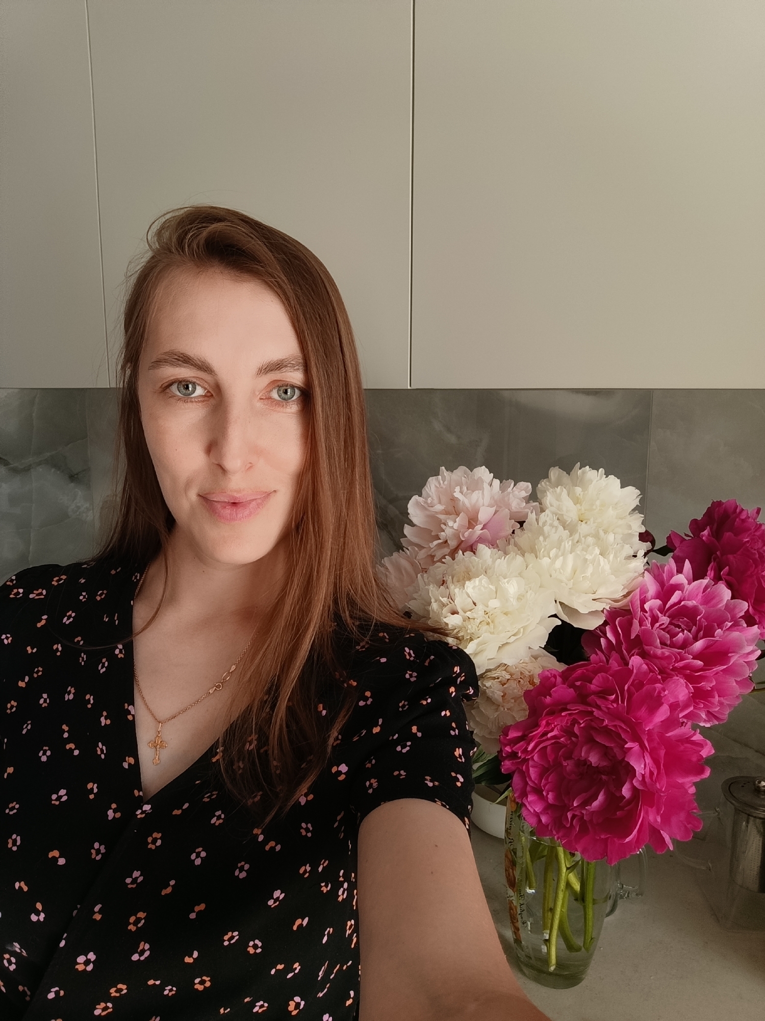 In a domestic indoor setting, a woman with shoulder-length auburn hair and light blue eyes is photographed from the chest up. She is wearing a black dress with a floral pattern of small pink and orange dots. Around her neck, she wears a thin gold chain with a cross pendant. To her right, a bouquet of peonies in shades of pink and white is arranged in a clear glass vase. The background consists of a neutral-toned wall, possibly kitchen cabinets, and a grey tiled backsplash. The overall lighting suggests an indoor environment. The image appears to be a selfie taken in a residence within the city of Chaikovsky, Russia.
