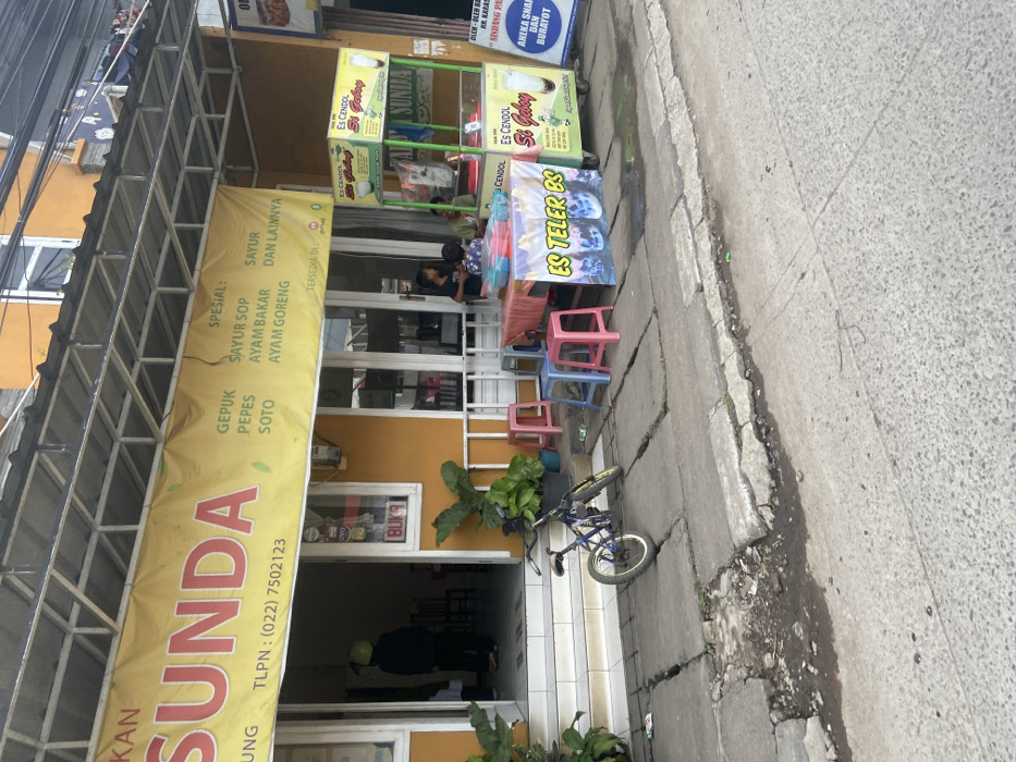 The image depicts a street scene in Bojongsoang, Indonesia, located at latitude -6.979 and longitude 107.634. In the foreground, there is a small, blue bicycle parked on the sidewalk. To the right of the bicycle, there is a small stand with a green frame. The stand is selling "Es Cendol Si Geloy" and "Es Teler Bs." There is also a sign that reads "Aneka Snack dan Buruyot." The stand has an opaque plastic container filled with a beverage and a clear plastic container with a red lid, which may contain a different beverage. Two people are sitting at a table with a red tablecloth. They are likely waiting for their orders.
In the background, there is a building with a large, yellow sign that reads "Sunda" in red letters. The sign lists various menu items in Indonesian, including gepuk, pepes, soto, sayur sop, ayam bakar, and ayam goreng. There is a person in the doorway of the building. It is not possible to see the gender of the person or what they are doing. The sidewalk is made of concrete blocks. The street is paved with asphalt. There are several green plants in pots in front of the building. The building is a yellow color and has a white awning. There is a white roof above the awning. The scene is a typical street scene in an urban area of Indonesia, with a mix of businesses and residential dwellings.