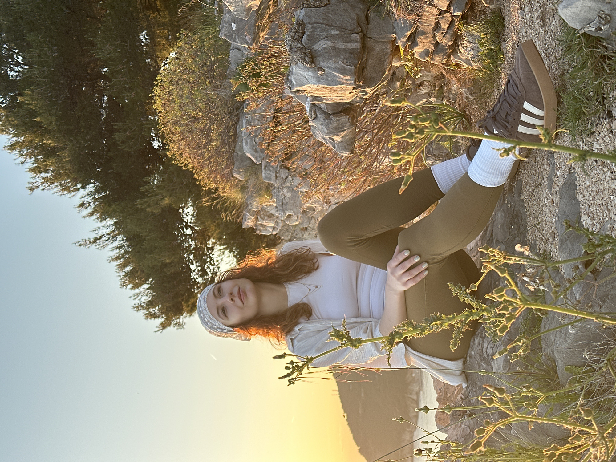 A person sits on a rock face. The person wears a white shirt, green pants, white socks, and grey sneakers with white stripes. A white patterned bandana is on the person’s head. The scene includes rocks, a tree, and a plant in the foreground. The scene is lit with warm light, and the sky is visible in the background. The location is Petrovac centar, Montenegro.