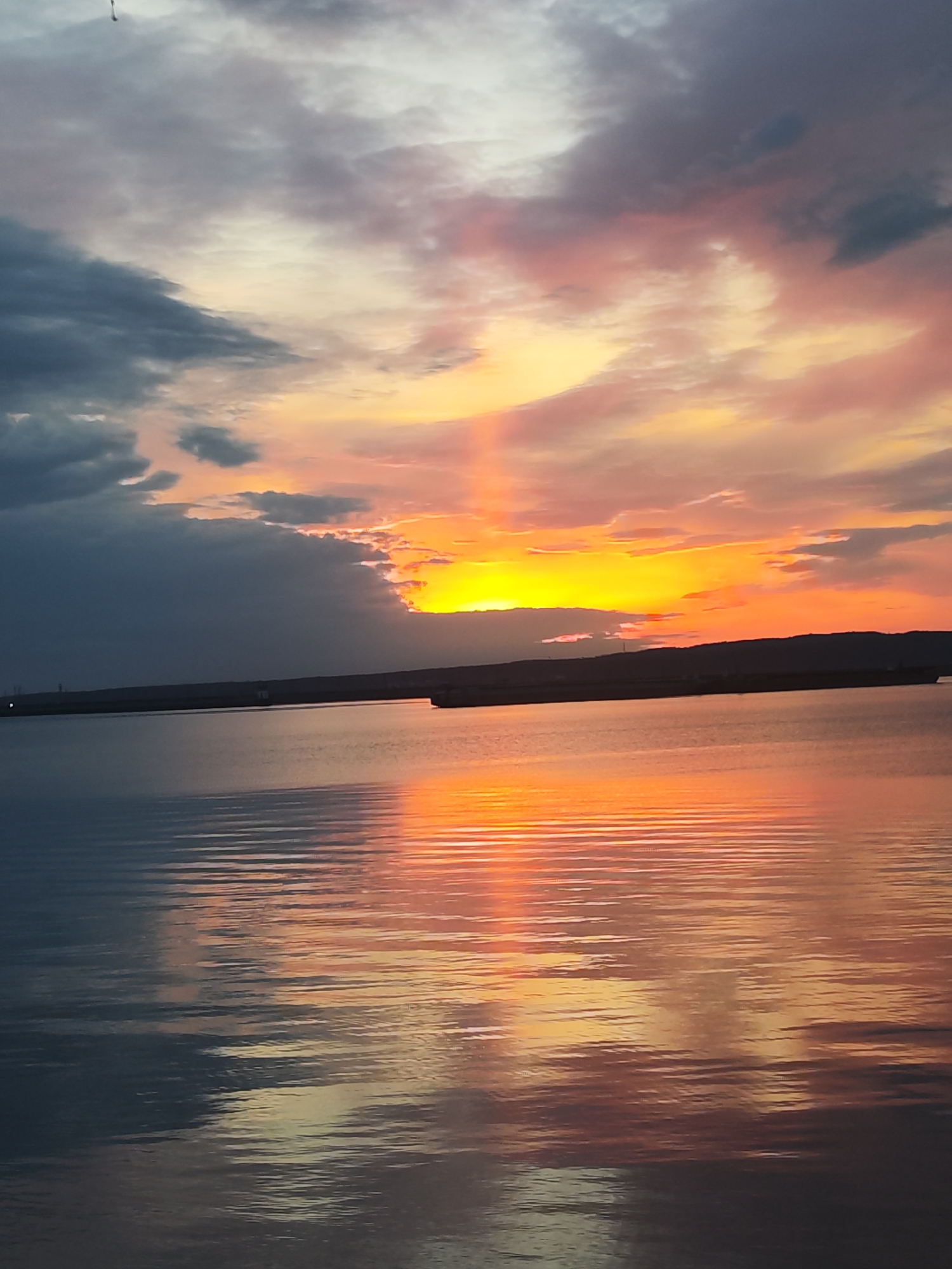 The image captures a serene sunset over the Kama River in Chaikovsky, Russia. The sky is filled with a mix of dark, grey clouds on the left transitioning to vibrant orange and pink hues towards the center and right, where the sun is setting. The sunlight breaks through a gap in the clouds, creating a vertical beam of light. The water reflects the colors of the sky, with horizontal ripples adding texture to the surface. In the distance, a dark, low-lying land mass is visible, silhouetted against the bright horizon.