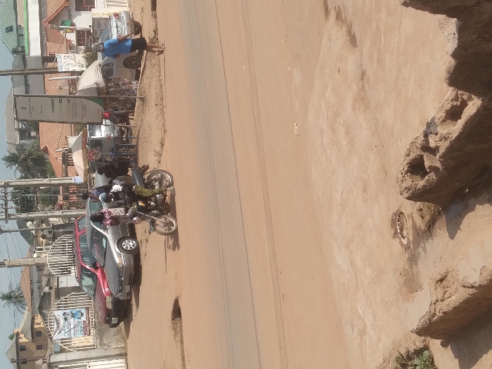 The image depicts a street scene in Agege, Nigeria. In the immediate foreground, an uneven, sandy-brown ground contains two irregular, dilapidated concrete or mud structures and a circular metal drain cover. The midground features a wide, unpaved reddish-brown dirt road with discernible tire tracks and a distinct pothole near the left curb. A person on a motorcycle, wearing a dark top, light-colored camouflage pants, and a dark helmet, is seen riding leftward on the road. Adjacent to the road, near a white metal gate, a red sedan and a silver sedan are parked. Three individuals stand near these vehicles: two females in patterned dresses and one male in a light-colored shirt. Further along the roadside on the right, several individuals are positioned under two light-colored market umbrellas, indicative of a vendor setup, with merchandise visible. Another silver sedan is parked near this area. A tall, white signpost and multiple utility poles with complex wiring intersect the upper portion of the scene. Various residential or commercial buildings with roofs of red, green, and brown hues are present in the background. On the far right, a female in a light blue top walks leftward, away from the camera. The scene is illuminated by what appears to be bright, diffused daylight.