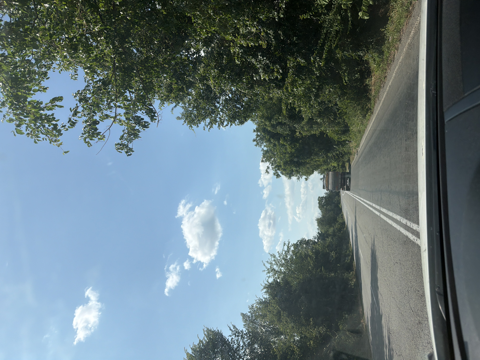 The image depicts a view from inside a vehicle looking out at a highway lined with lush green trees under a bright blue sky with scattered white clouds. The road is asphalt and has white double lines marking its center. In the distance, a truck is visible driving on the road. The scene suggests a sunny day, possibly in the late morning or early afternoon, given the quality of light and the shadows cast. The presence of dense foliage on either side of the road indicates a rural or semi-rural setting, possibly a forest or park area. The perspective from within the vehicle, with the edge of the vehicle's interior visible on the right side of the frame, suggests the photograph was taken while traveling. There is no visible text in the image.