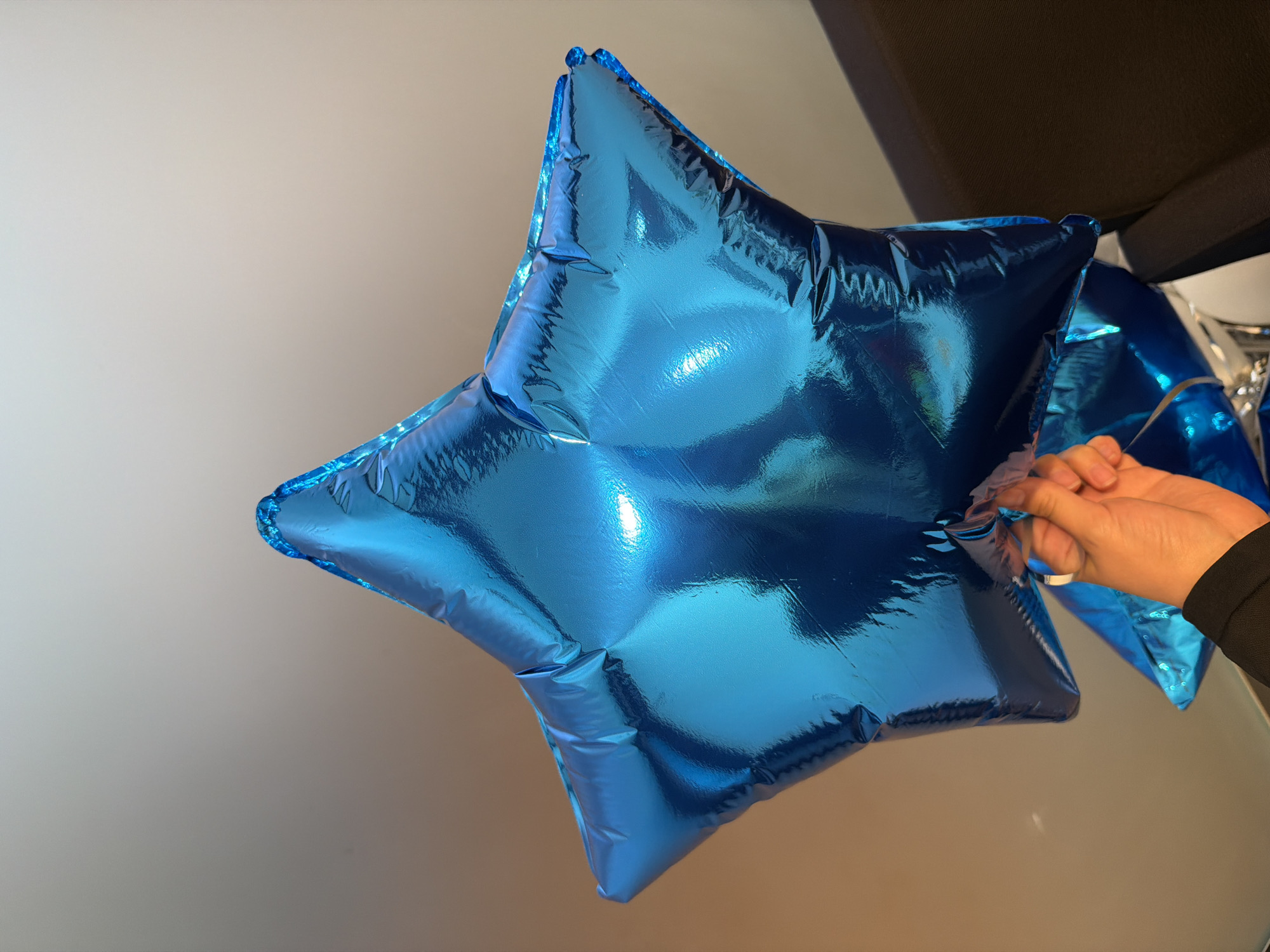 The image shows a close-up of a shiny blue star-shaped balloon. A hand is visible, holding the balloon and appearing to be preparing it for inflation or deflation, as the edge of the balloon material is being manipulated. Another blue balloon is visible in the background, suggesting a festive or celebratory context. The lighting is bright, creating reflections on the metallic surface of the balloon. The background is a plain, light-colored wall, providing a simple backdrop that emphasizes the balloon. The scene appears to be indoors, possibly in a home or event space. No text is visible in the image.