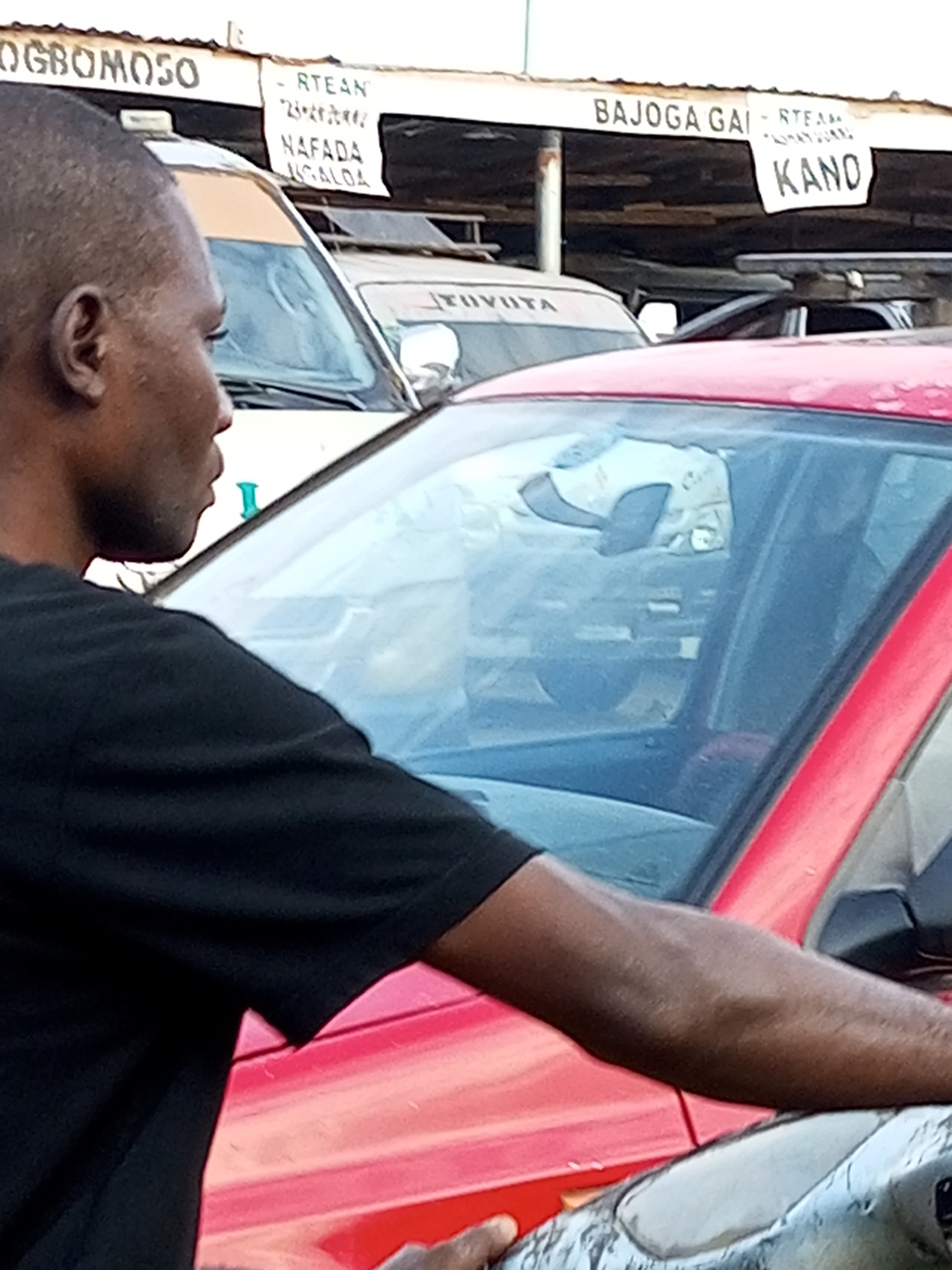 The image depicts a street scene in Gombe, Nigeria, with a focus on vehicles and signage. In the foreground, a man wearing a black t-shirt is visible from the side, with his arm resting on the hood of a red car. His left arm and shoulder are prominent. Behind him, a white vehicle, possibly a commercial bus, is parked. The side of this white vehicle has "TOYOTA" written on it. Above the vehicles, there is a structure with various signs. One sign, partially visible on the left, reads "OGBOMOSO". Another sign to the right displays "BAJOGA GAI - RTEAN" and below it, "KANO". There are also other smaller signs with text that is difficult to discern. The overall environment suggests a busy roadside area, likely a transportation hub or market, given the presence of commercial vehicles and route signage. The lighting indicates it is daytime.