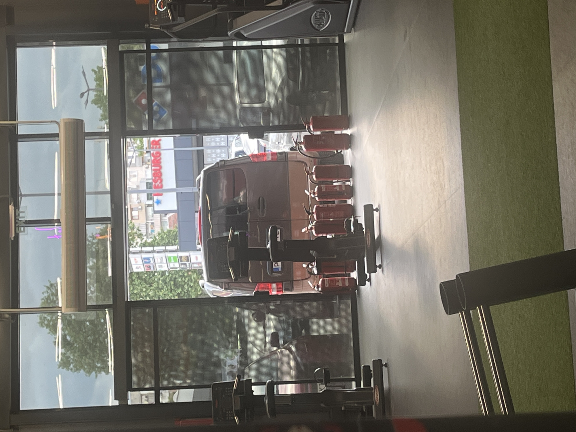 The image shows the interior of a gym in Plovdiv, Bulgaria, with a view through the windows towards a street scene. Outside, a commercial building with visible signs for "Hesburger" and "Domino's Pizza" can be seen. A silver van is parked outside. Inside the gym, various exercise machines are visible, and multiple red fire extinguishers are mounted on a stand. The floor is light-colored, and there is a section of green turf on the right side of the frame. The overall lighting is bright, suggesting daytime.