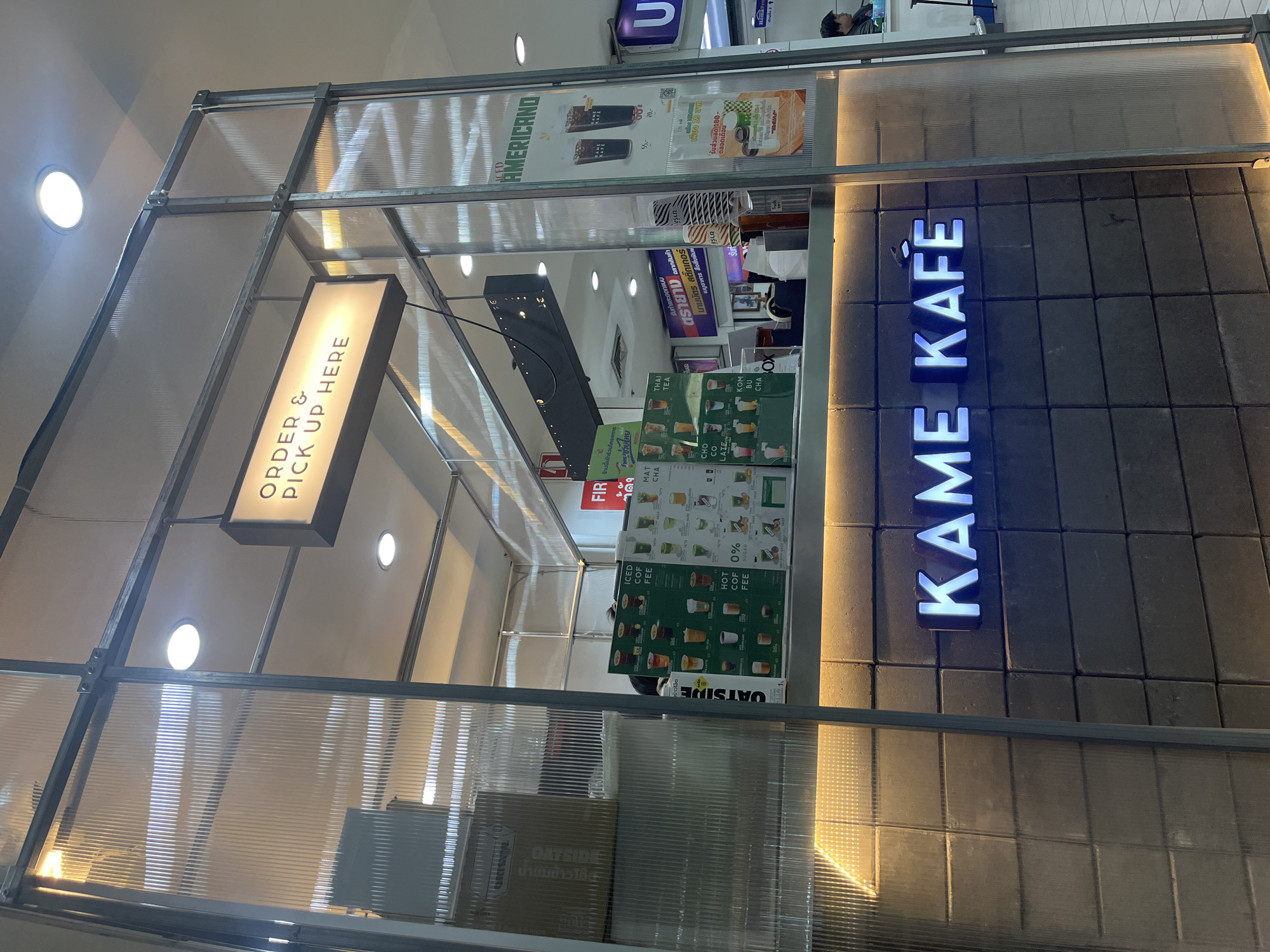 The image shows the exterior of a coffee shop called "KAME KAFE" in Nong Pa Khrang, Thailand. The shop is enclosed by a structure made of metal framing and translucent corrugated panels. Inside, a menu board displays various coffee and tea options, including iced and hot coffee, Thai tea, matcha, and chocolate latte. Prices are visible, such as "55.-" and "70.-". A sign above the counter reads "ORDER & PICK UP HERE".
The lighting suggests it is daytime, with warm light coming from inside the shop and the ceiling lights. The overall aesthetic is modern and minimalist, with a dark tiled wall behind the "KAME KAFE" sign which is illuminated with blue neon lights. The scene appears to be set in a public area, possibly a mall or market, given the surrounding environment and the presence of other signage in the background. One person is partially visible behind the counter, appearing to be a staff member. The atmosphere seems calm and inviting, typical of a casual coffee shop setting.