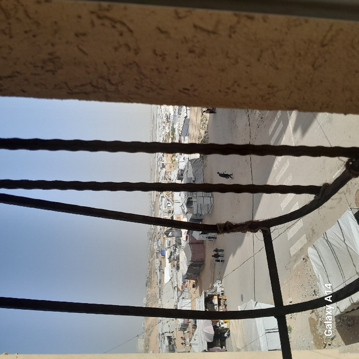 A view through a window with a barred frame reveals a sprawling refugee camp. The tents and makeshift structures are densely packed across a dusty landscape. A road runs through the camp, with several people walking along it. The sky is hazy, suggesting a dry or polluted environment. The window frame is visible, adding depth to the scene.