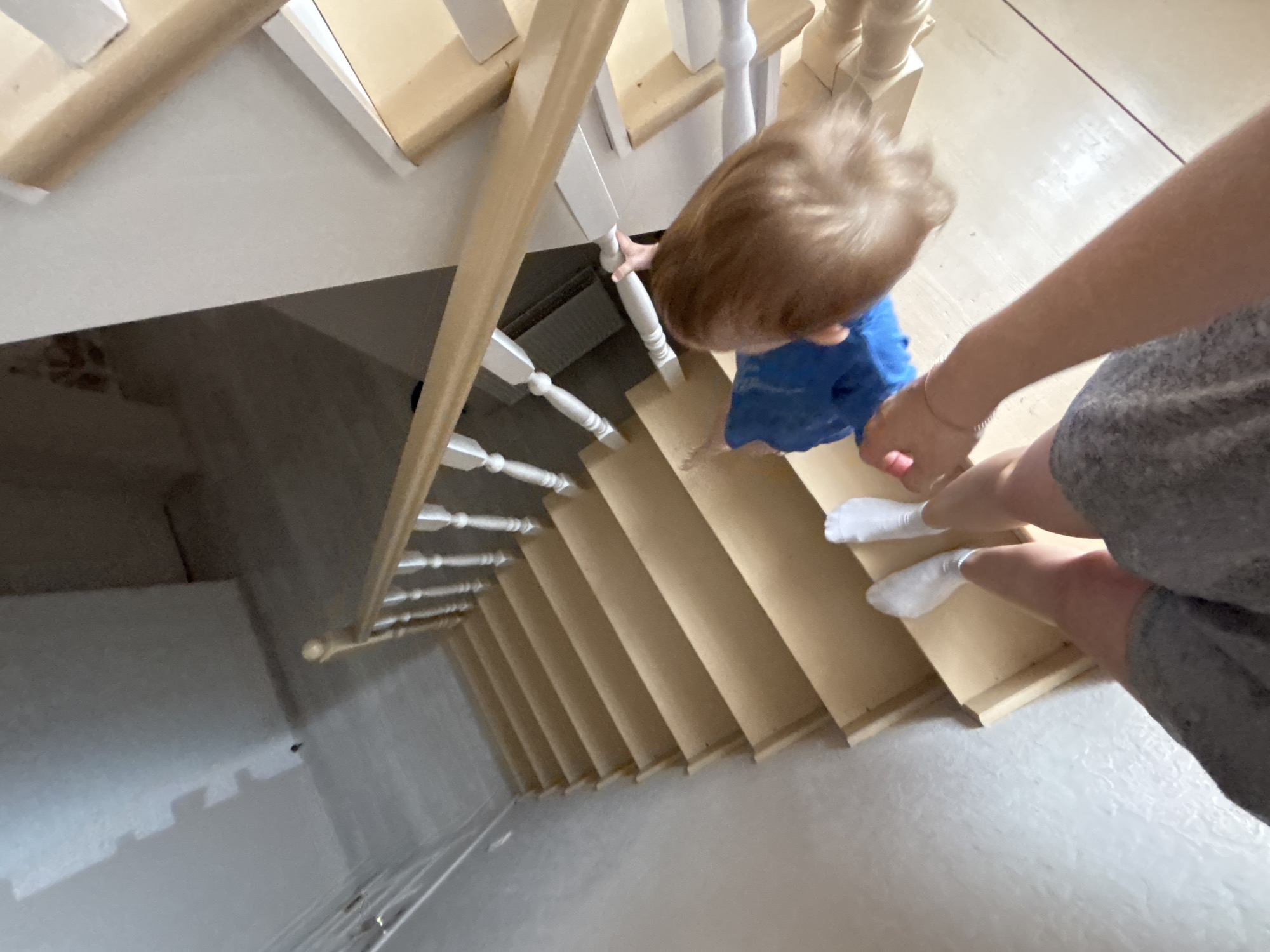 A high-angle view shows a child and an adult descending a wooden staircase. The child, wearing a blue shirt, holds the white railing on the left. The adult, wearing a gray outfit and white socks, follows closely behind, possibly assisting the child. The stairs are made of light-colored wood and have white railings. The walls beside the stairs are partially painted, with visible signs of renovation. The staircase is located inside a building in Ферма, Russia.