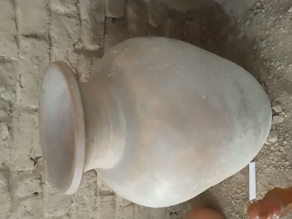 A large, unglazed clay amphora sits on a dusty floor in front of a rough brick wall. The amphora is light brown and has a rounded body that tapers to a narrow neck and a wide, flared opening. The surface of the amphora is uneven and shows some smudges and dust. The brick wall behind it is made of rough, rectangular bricks that are unevenly laid and appear weathered. The floor is covered with dirt and small pebbles. In the lower left corner of the image, part of a small, reddish-brown clay object is visible, possibly a fragment or a small statue. A white label with text is also visible on the floor in front of the amphora. The overall scene suggests an archaeological or historical exhibit. This scene is located in the city of Angren, Uzbekistan.