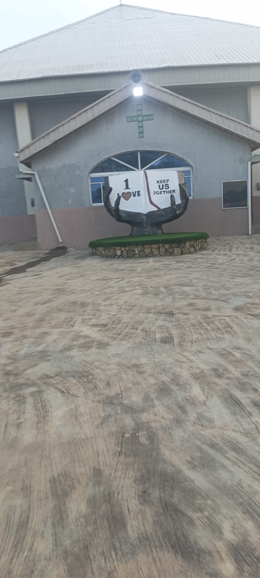 A gray concrete-paved courtyard leads to a gray, one-story building, likely a church, in Jos, Nigeria. The building has a gray metal roof and a symmetrical gable with a green cross centered on it. A speaker is mounted above the cross. A large, sculptural monument depicting open hands holding an open book stands in front of the building. The book displays the words "1 Love" and "Keep Us 2gether." The monument is surrounded by a small, circular patch of artificial green turf enclosed by small, tan stones. A drainpipe runs down the side of the building. A small window is visible to the right of the monument. The sky above is overcast.