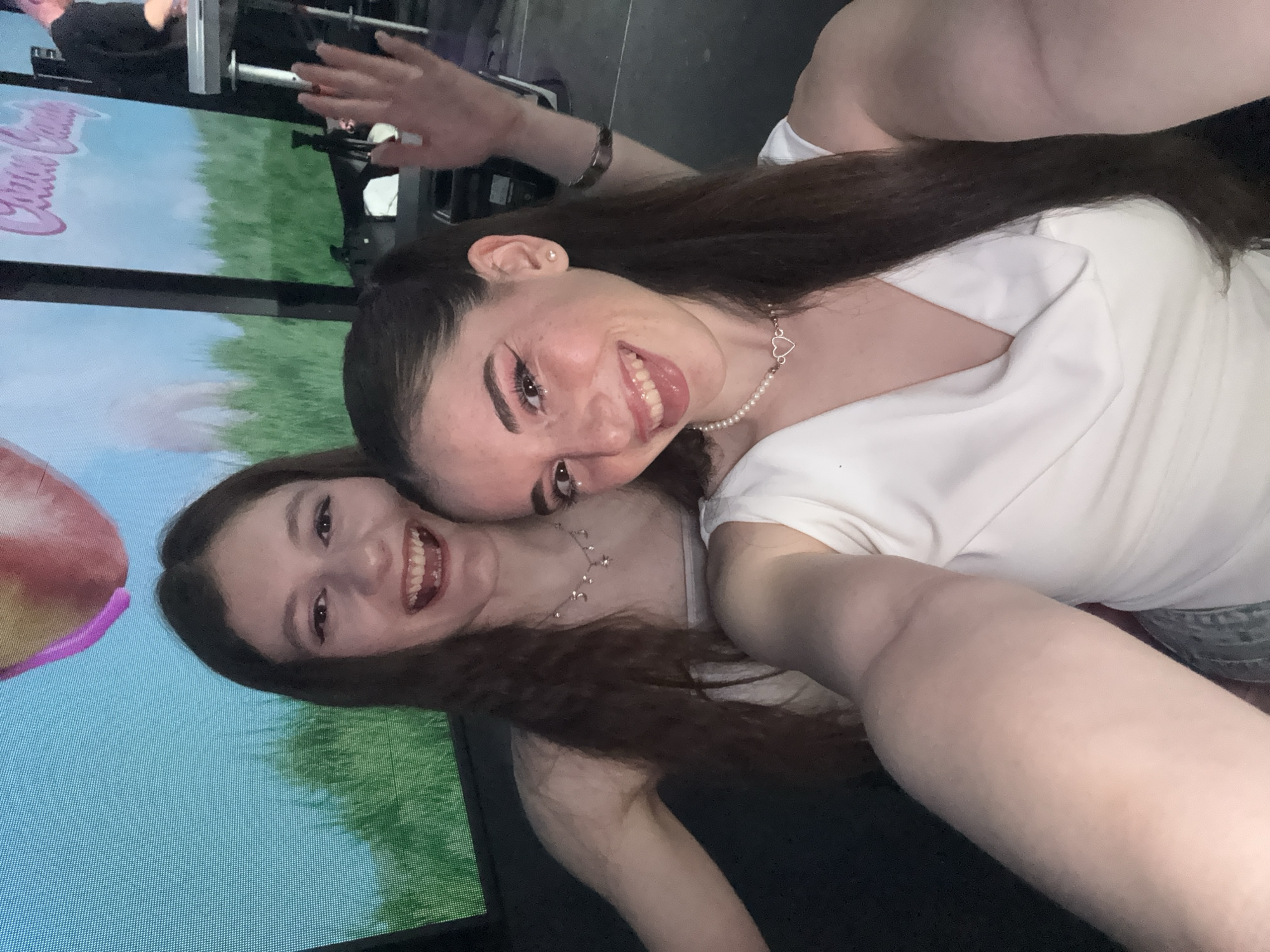 Two women are posing for a selfie. One woman is wearing a white top and a pearl necklace with a heart pendant. She is sticking her tongue out. The other woman has on a delicate necklace. Both women have long brown hair. A screen in the background displays a landscape. The location is not identifiable as Plovdiv, Bulgaria, from the given image.