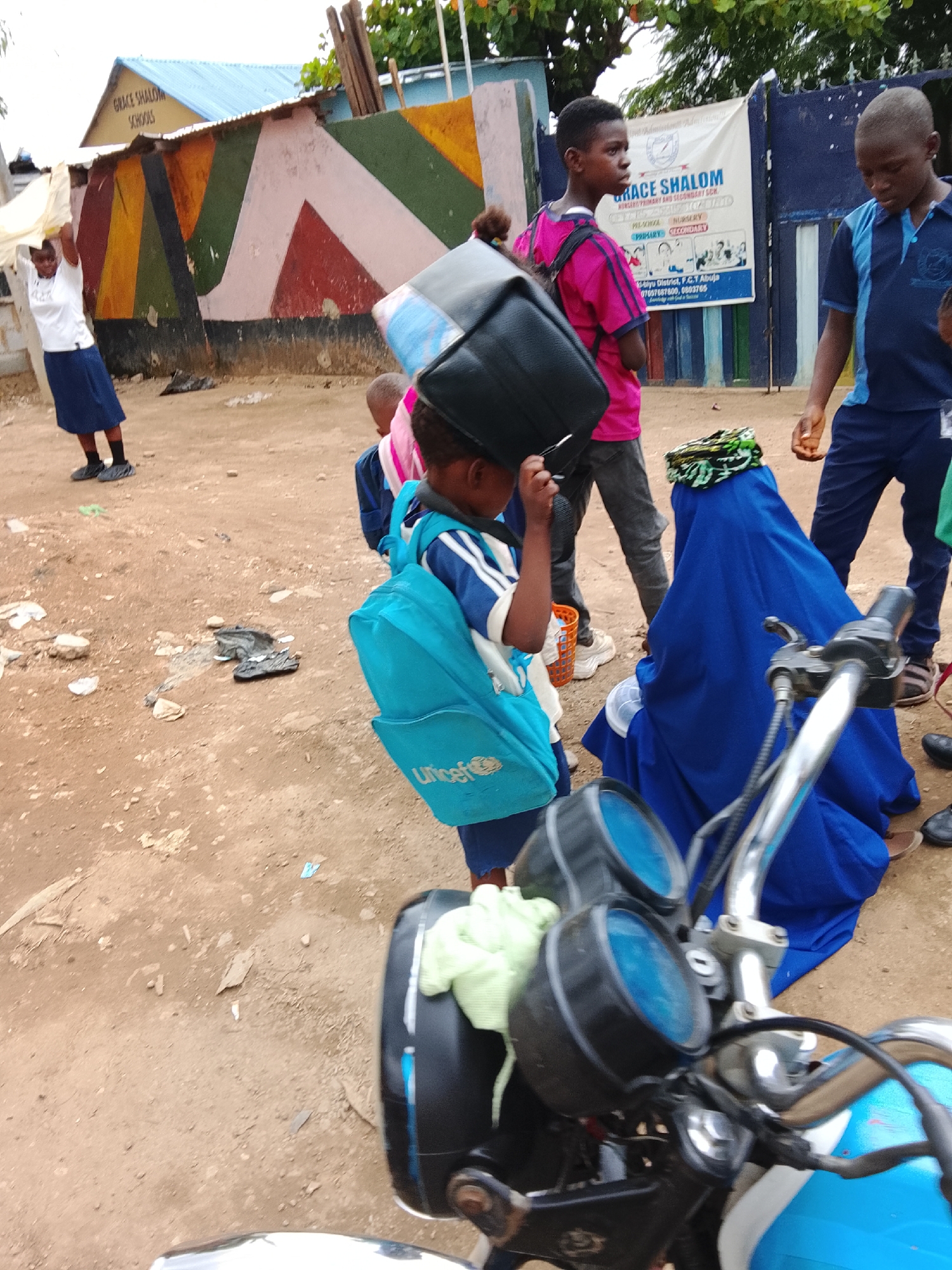The image depicts a scene at the Grace Shalom Schools, located in the Gaduwa District, F.C.T. Abuja, Nigeria. Several people, mostly children in school uniforms, are present. A boy with a blue UNICEF backpack is carrying a black object on his head. A woman in a blue burqa sits nearby. A motorcycle is parked in the foreground, partially obscuring the view. In the background, a building with a sign reading "Grace Shalom Schools" and a colorful wall is visible. Some litter is scattered on the ground.