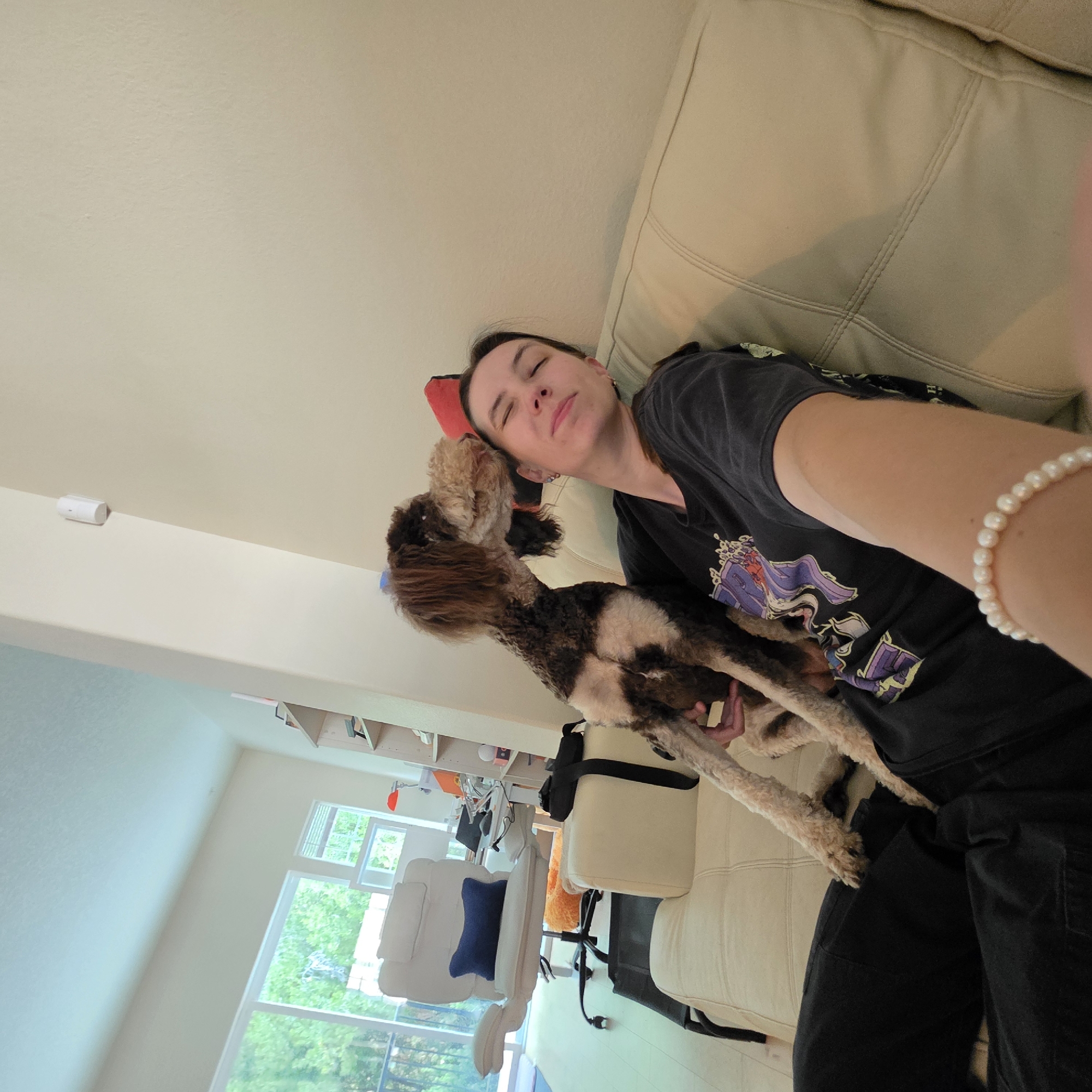 In a domestic indoor setting in Portland, Oregon, a woman is seated on a cream-colored couch. She is wearing a dark t-shirt and black pants, with a pearl bracelet on her right wrist. A medium-sized dog, with brown and white fur, is positioned on her lap and leaning towards her face, appearing to be licking or nuzzling her cheek. The woman has her eyes closed and a content expression. Behind them, a wide window reveals a view of lush green trees. To the left of the woman, a white office chair with a blue cushion is visible, along with a desk and shelves in the background, suggesting a living or work area within a home. The wall is a neutral, light color.